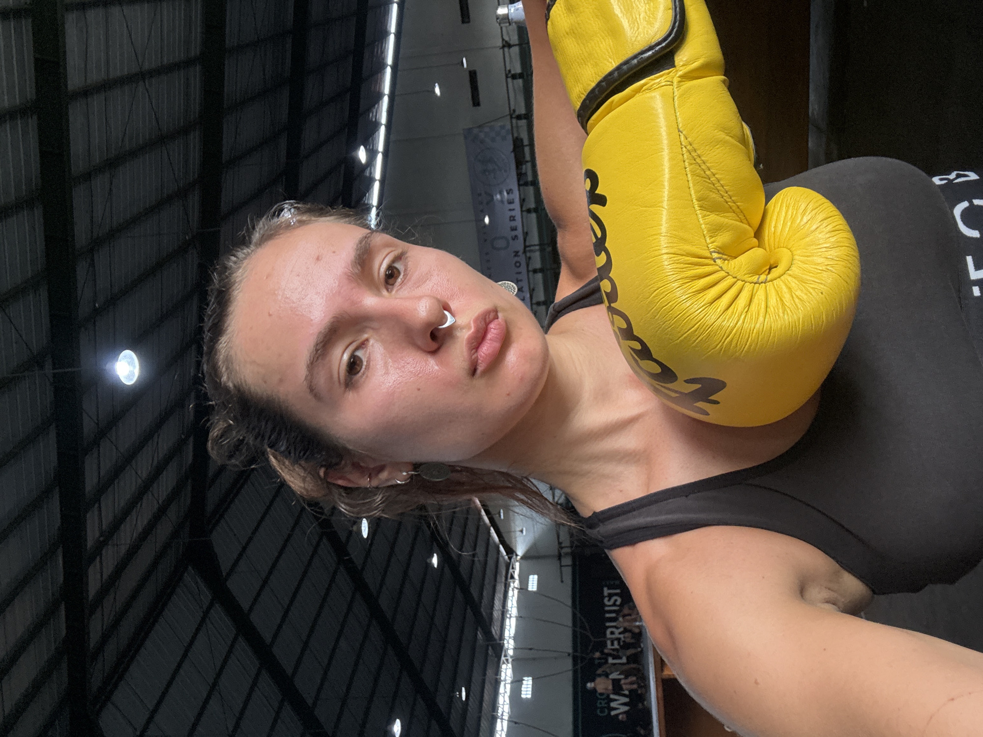 A selfie shot from a low angle features a woman in a black sports bra and leggings, holding a yellow boxing glove near her face. The woman is fair-skinned and has light brown hair pulled back from her face. She wears a nose ring and earrings. The boxing glove is bright yellow with black accents and writing. The background includes the interior of a large, industrial-style building with a dark ceiling supported by metal beams and bright lights. A banner in the background is partially visible and features text including the words "WANDERLUST". Based on the banner and the general appearance of the location, this image was likely taken at the "Crossfit Wanderlust" gym in Tibubeneng, Indonesia.
