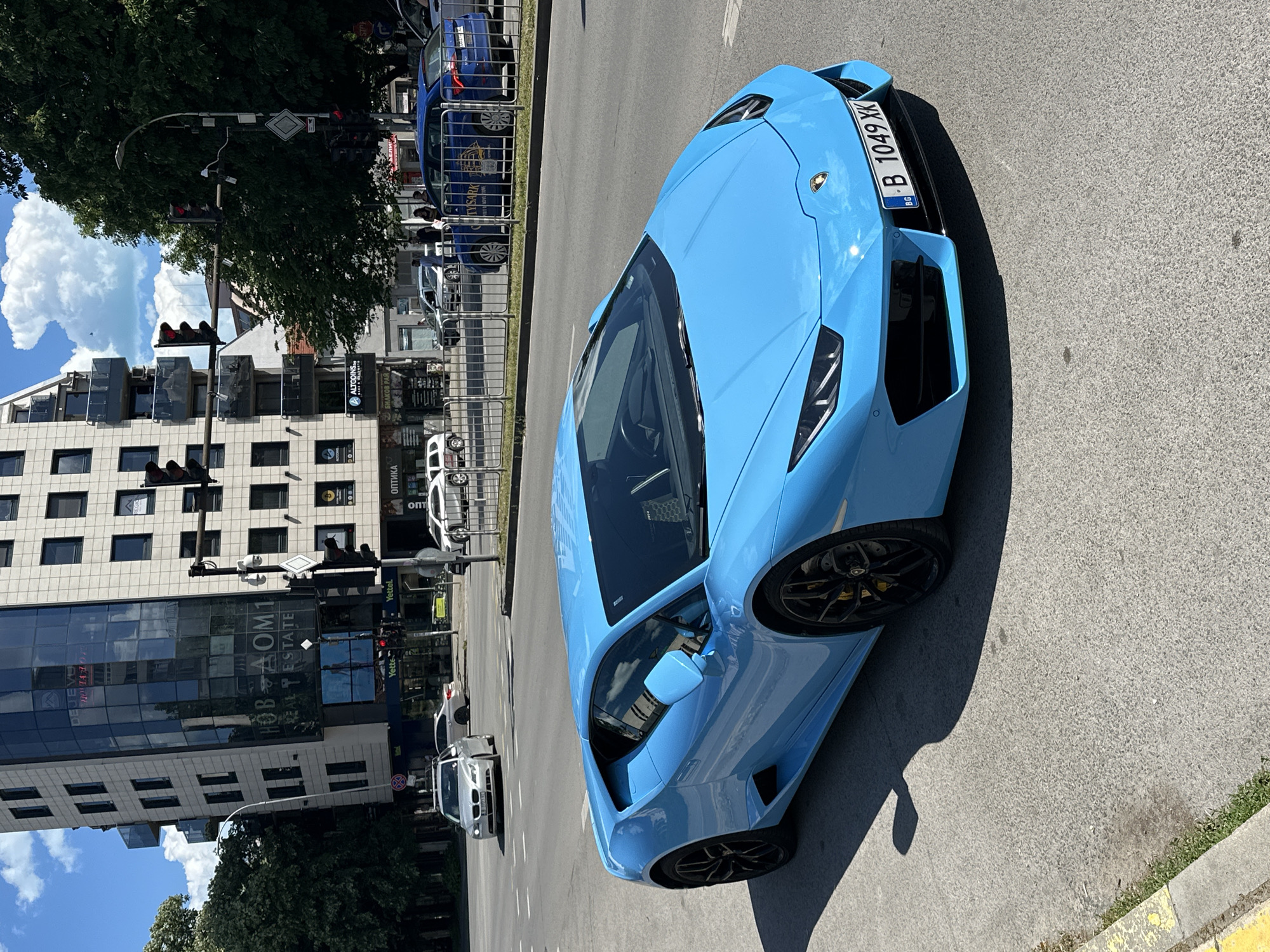 The image shows a light blue Lamborghini Huracan parked on a street in Varna, Bulgaria. The license plate reads "B 1049 XK". Behind the car, there is a building with "НОВ ДОМ 1 REAL ESTATE" visible on its facade. Traffic lights and other vehicles are also present on the street. There are trees on both sides of the street, and the sky is visible with some clouds.