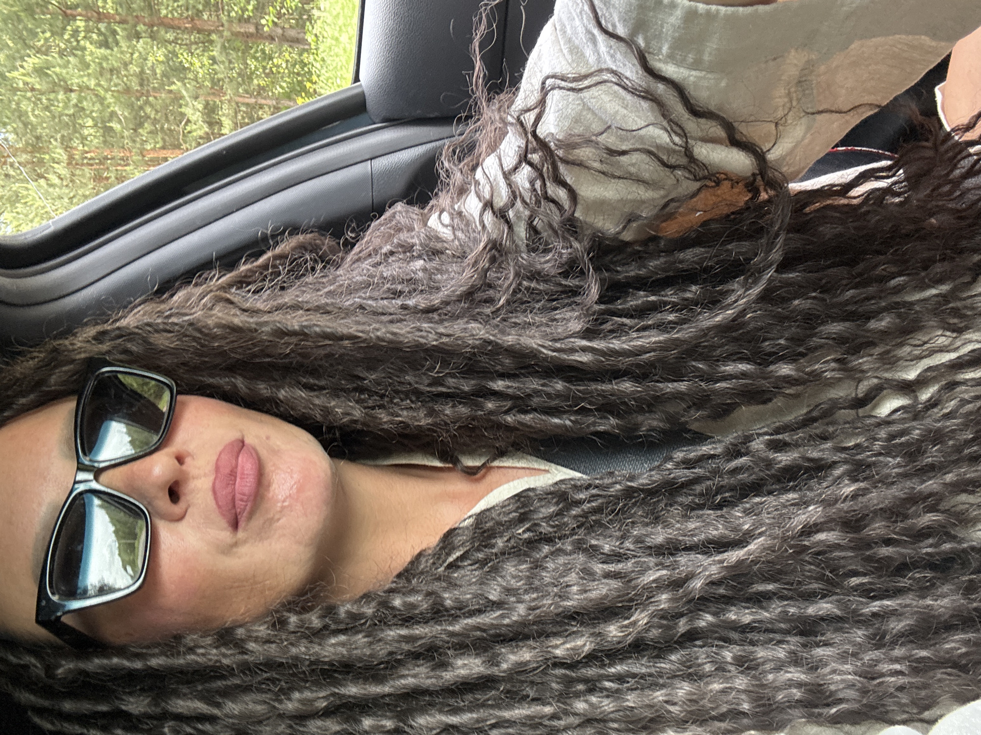 This image features a person, likely female, captured inside a car in Chaikovsky, Russia. The subject's face is visible in the lower left portion of the frame, oriented sideways with her head resting. She has long, dark, tightly curled or textured hair that flows across the car interior, dominating a significant part of the image.
The person is wearing dark-framed sunglasses, reflecting the bright green foliage outside the window. Her lips are visible with a soft pink lipstick, and her expression appears calm and neutral, suggesting relaxation or rest. She is dressed in a light-colored top, possibly white or off-white.
The scene is set inside a vehicle, likely a passenger car, with a black door panel and a car window visible. Outside the window, a dense background of green trees and foliage indicates a natural, possibly forested or rural environment. The bright natural light suggests it is daytime, and the weather appears clear and sunny. No specific activity beyond resting or observing the surroundings is depicted. There is no visible text in the image.