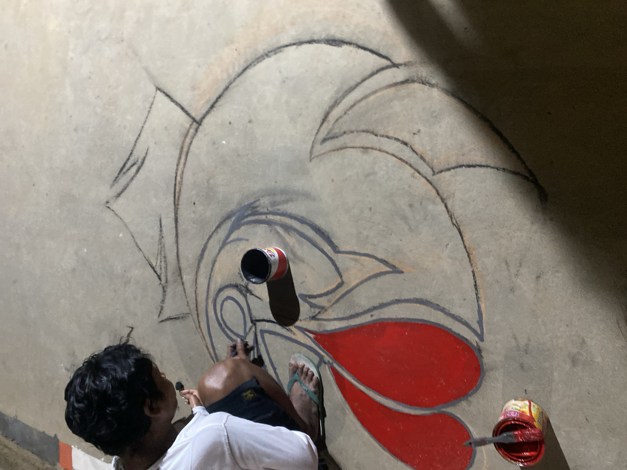 An overhead, high-angle medium shot captures a person kneeling on a concrete surface, engaged in the creation of a large chalk drawing. The person, seen from the back and right side, has dark hair and is wearing a white t-shirt and dark shorts. They are actively working on the drawing with their right hand, holding a small object, likely a chalk stick or brush. To their left, a can of red paint sits open with a brush inside, and further down, two large red shapes have already been painted as part of the design. The drawing itself appears to be a stylized representation of a large, circular form with intricate lines and curves, outlined in dark chalk. The concrete surface shows signs of wear and is slightly uneven. The lighting casts shadows, indicating it may be evening or an indoor setting with artificial light. The background is a continuation of the concrete surface, with some indistinct lines or smudges. The overall scene suggests an artistic activity taking place on the ground. The location is Unknown, Unknown.