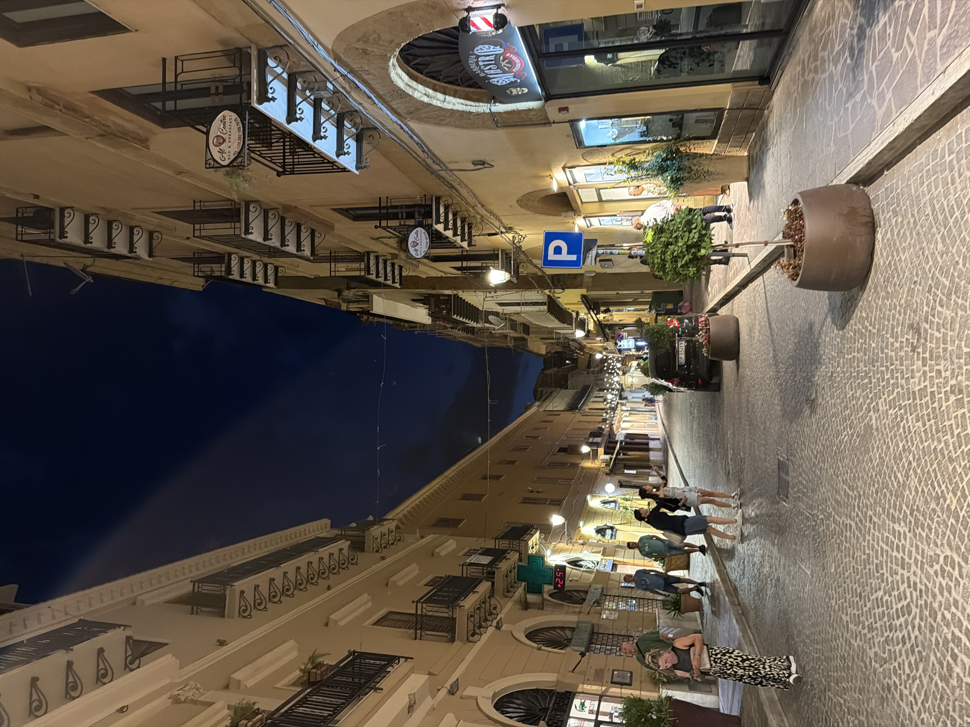 A street scene in Agrigento, Italy, at night, features buildings lining both sides of a cobblestone street. On the left, a building with ornate balconies and decorative trim is illuminated by streetlights. A green cross sign with a temperature display of +25 is visible. On the right, another building showcases a barber shop sign with a red and white striped pole. Several people are walking on the street, including a woman in a patterned dress on the left and a group of three individuals further down the street. A parked car is visible on the right side of the street. The sky above is dark, indicating nighttime.