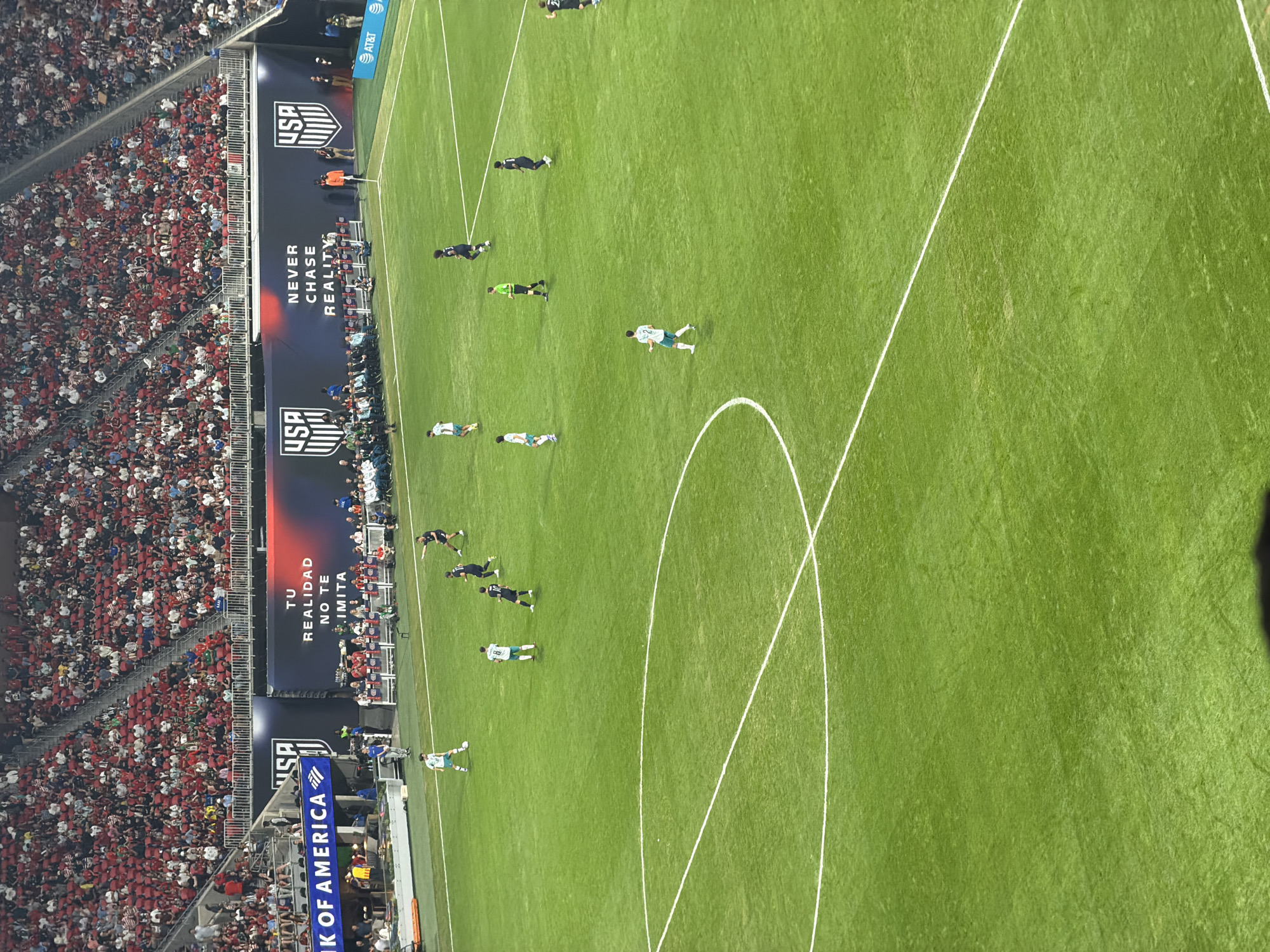 The image shows a soccer game in progress at a stadium, likely in Atlanta, United States of America, based on the USA and Bank of America advertisements. Several players wearing dark jerseys are positioned on the green field along with a couple of players in light jerseys. The field is marked with white lines. Spectators are seated in a crowded stadium with red colors dominating the crowd, sitting on the stadium steps. Advertisements and banners are visible along the side of the field. The AT&T logo is seen. The scene appears to be captured from a high vantage point.