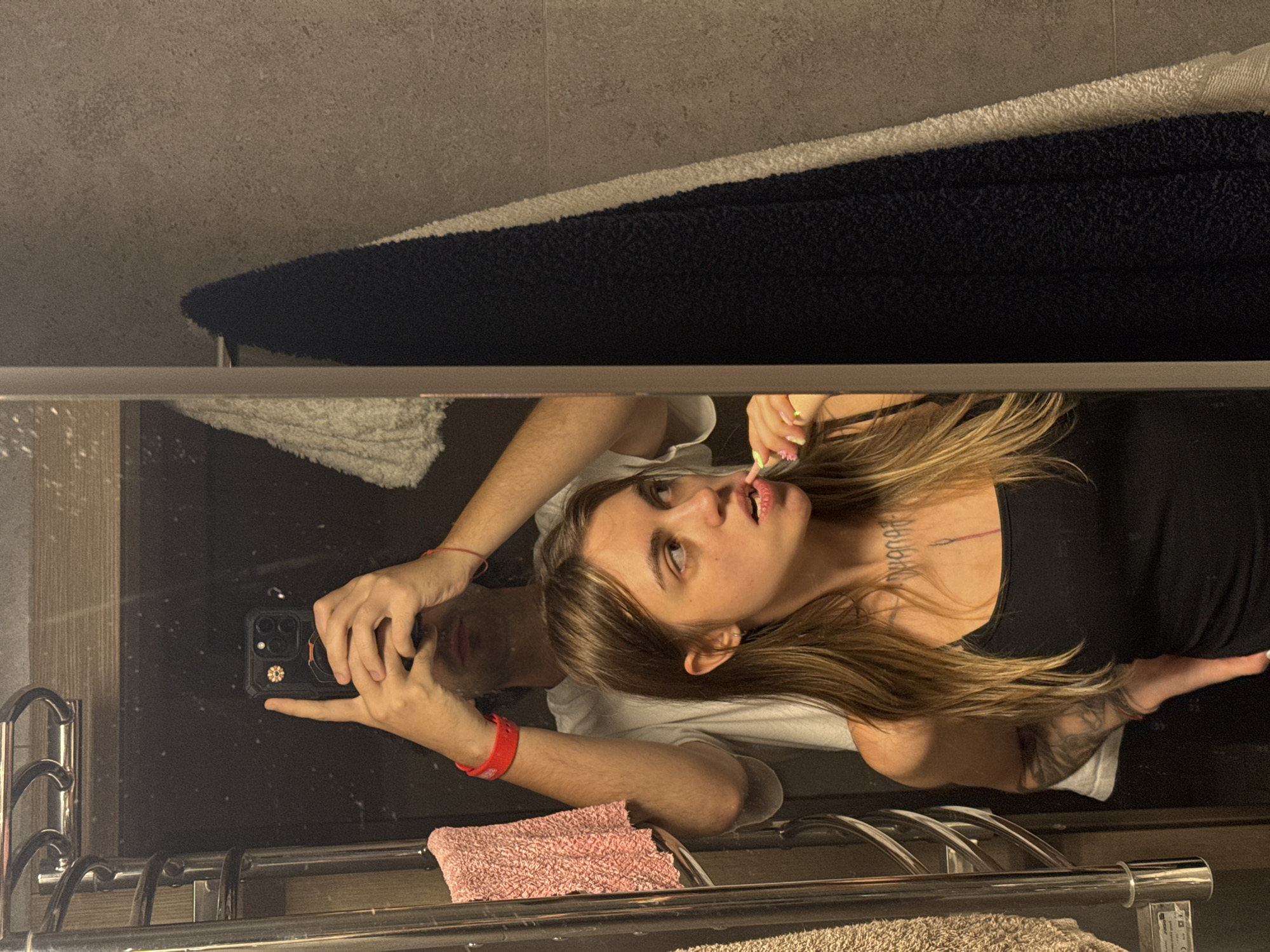 In a bathroom setting, a young woman with long, brown hair is brushing her teeth while looking to her right. She is wearing a black tank top. Behind her, a man is taking a selfie in the mirror, holding a phone in his hands. He is wearing a white t-shirt and has red bracelets on both wrists. The bathroom features a towel rack with a pink towel, and two other towels hanging on the wall, one black and one white. The walls are tiled with a textured, grey material.