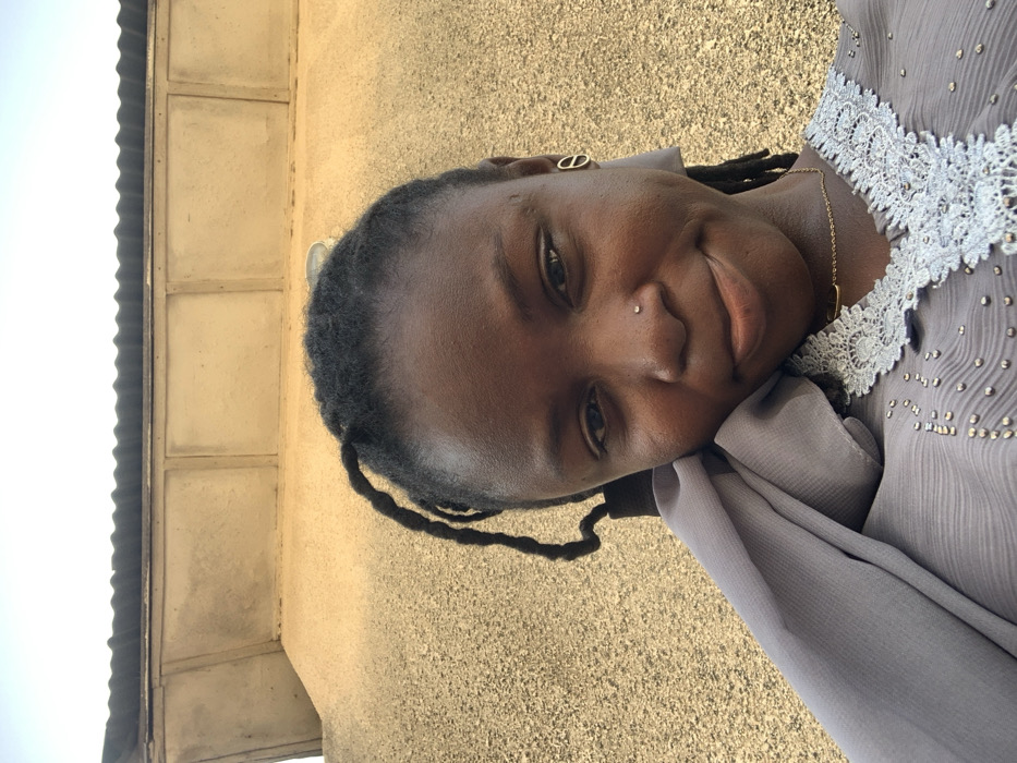 The image displays a young Black female outdoors in the city of Bojude, Nigeria. She is positioned facing the camera, exhibiting a slight smile. Her hair is dark, styled in dreadlocks or braids. A small, clear stud pierces her left nostril. She wears a distinct gold-toned oval-shaped earring on her right ear and a gold-colored chain necklace with a small horizontal bar pendant. Her attire includes a gray garment with a light-colored lace V-neckline and scattered shiny embellishments on the chest. A section of matching gray fabric is draped over her left shoulder. The background consists of a light brown, textured building wall featuring rectangular recessed panels, surmounted by a dark corrugated roofline. A clear, light blue sky is visible above the roof. The scene is illuminated by natural daylight.