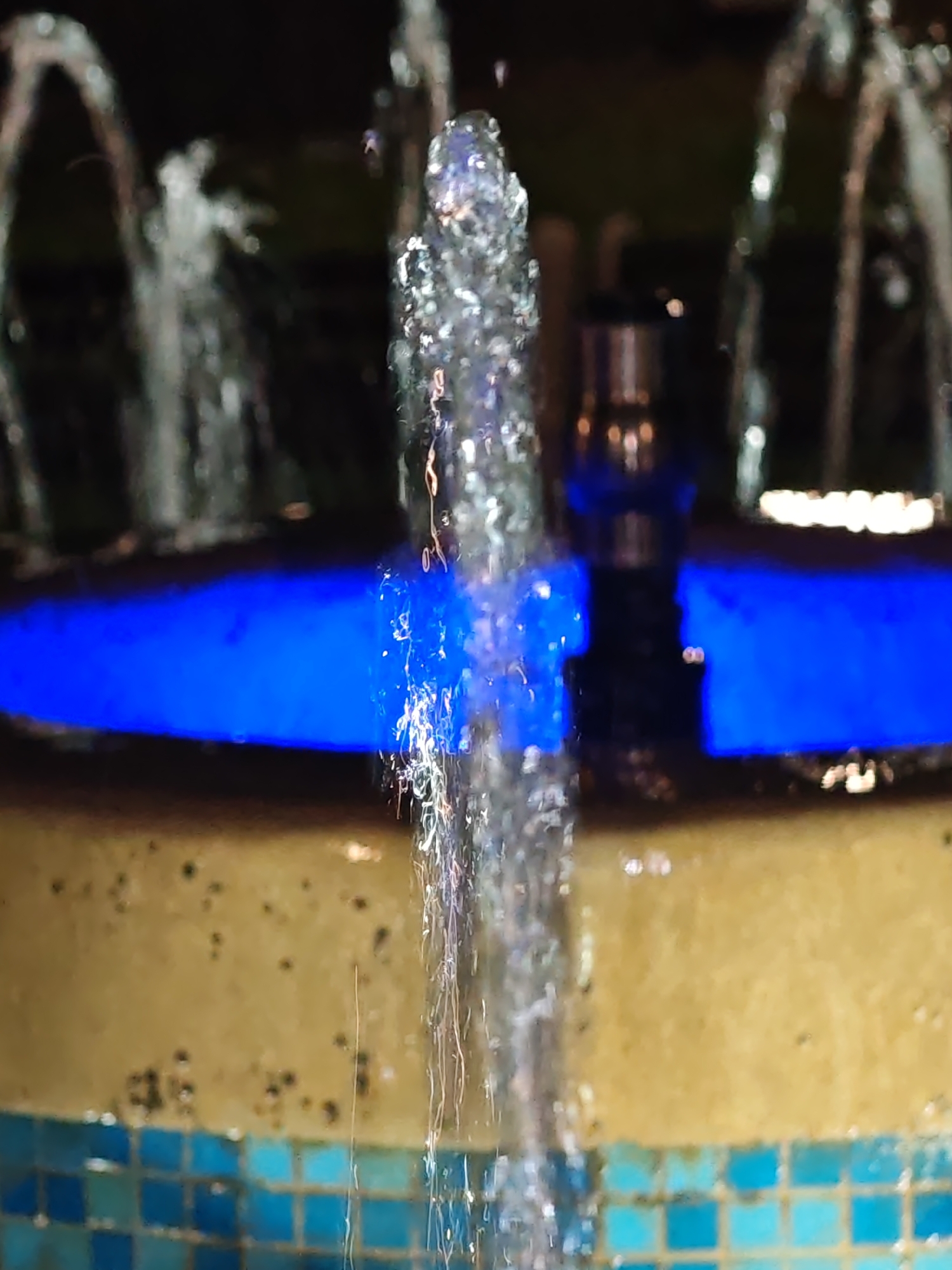 The scene captures a section of a water fountain at night in Veliki Preslav, Bulgaria. A central, vertical stream of water, appearing as illuminated droplets and thin filaments, rises in the foreground. Additional, out-of-focus water jets are discernible as blurred arcs and vertical streams in the dark background. The water is illuminated by a strong, artificial blue light source originating from within the fountain structure, casting a distinct blue glow on a horizontal surface in the mid-ground. A partially visible, reflective metallic cylindrical component is situated in the mid-ground, right of center. The lower portion of the fountain structure features a band of small, square mosaic tiles in varied shades of blue and teal. Above the tiles, a wider, yellowish-brown textured surface displays numerous small, dark specks. No persons are present.