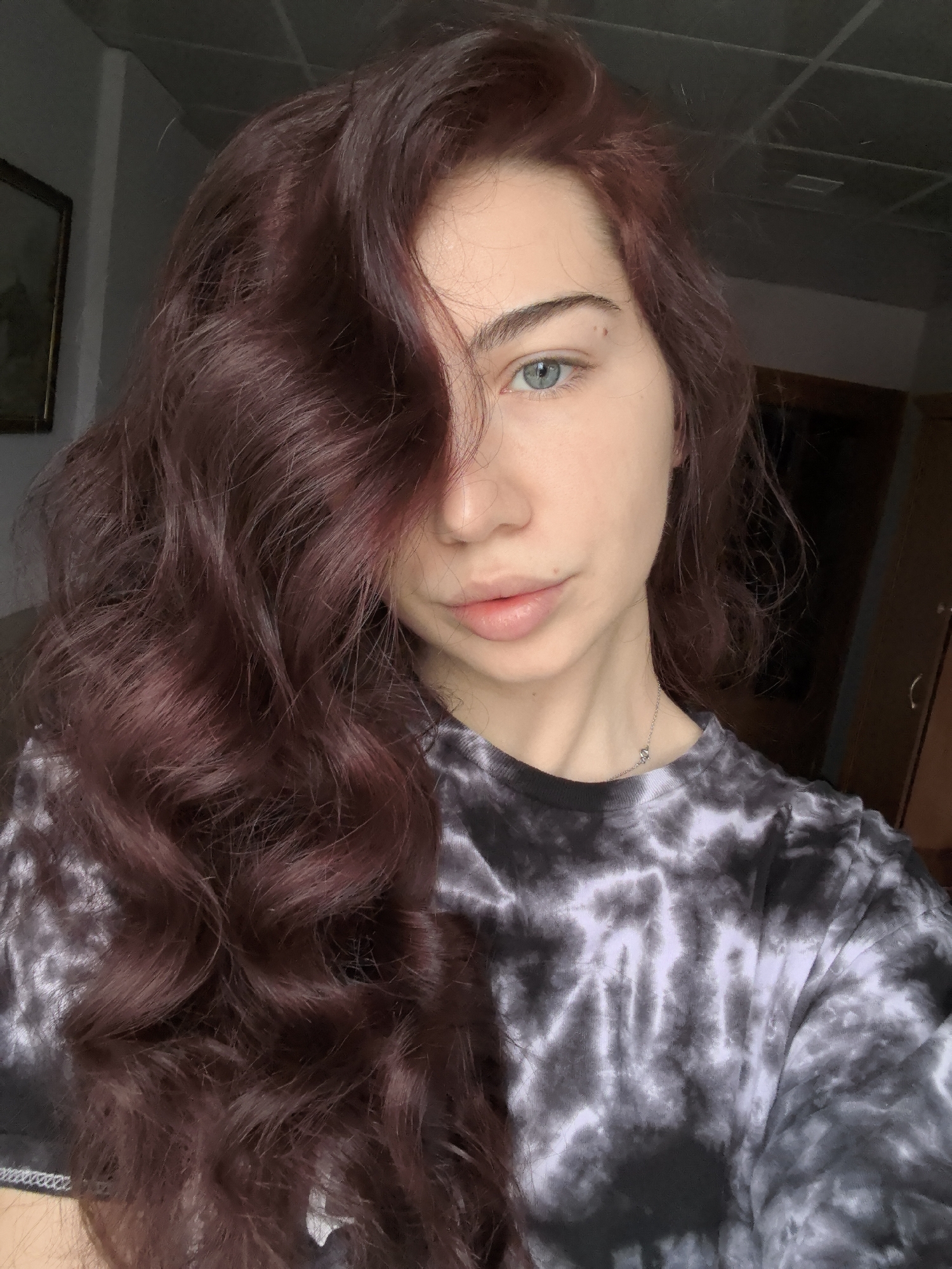A single female individual is depicted in an indoor setting, identified as being within the city of Stara Zagora, Bulgaria. The subject, positioned centrally, is a fair-skinned individual with long, wavy, reddish-brown hair, largely obscuring the left side of the face. The right eye, blue in color, is visible and directed towards the camera. A small dark mole is present on the right cheek, inferolateral to the right eye. The nose is straight, and the lips are full and naturally colored, slightly parted. The subject wears a black and white tie-dye long-sleeved crewneck shirt and a thin silver-colored necklace with a small, indistinct pendant. The action captured is consistent with a self-portrait.
The background elements are out of focus. To the subject's left, a light-colored wall features a portion of a dark-framed picture. Above and slightly to the right of the subject, a dark ceiling with a grid-like pattern of recessed squares or panels is visible, with a small rectangular light fixture or vent within one panel. To the subject's right, a dark brown vertical surface, possibly a wardrobe or door, is partially visible. Lighting appears to originate from an unseen source to the front-left, casting subtle shadows.