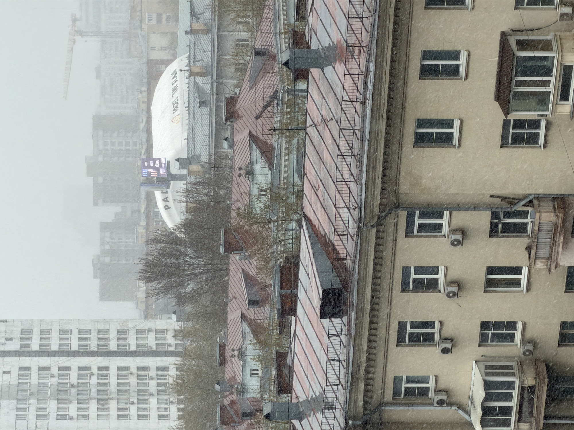 The image shows a cityscape in Moscow, Russia, during a snowfall. In the foreground is a residential building with windows, some of which have air conditioning units. Adjacent to this building are rooftops with metal fire escapes. In the background is a hazy view of other buildings, including a building with a curved, white roof with the words "Padel Time" on it, and a construction crane. The overall environment suggests an urban setting experiencing winter weather conditions.