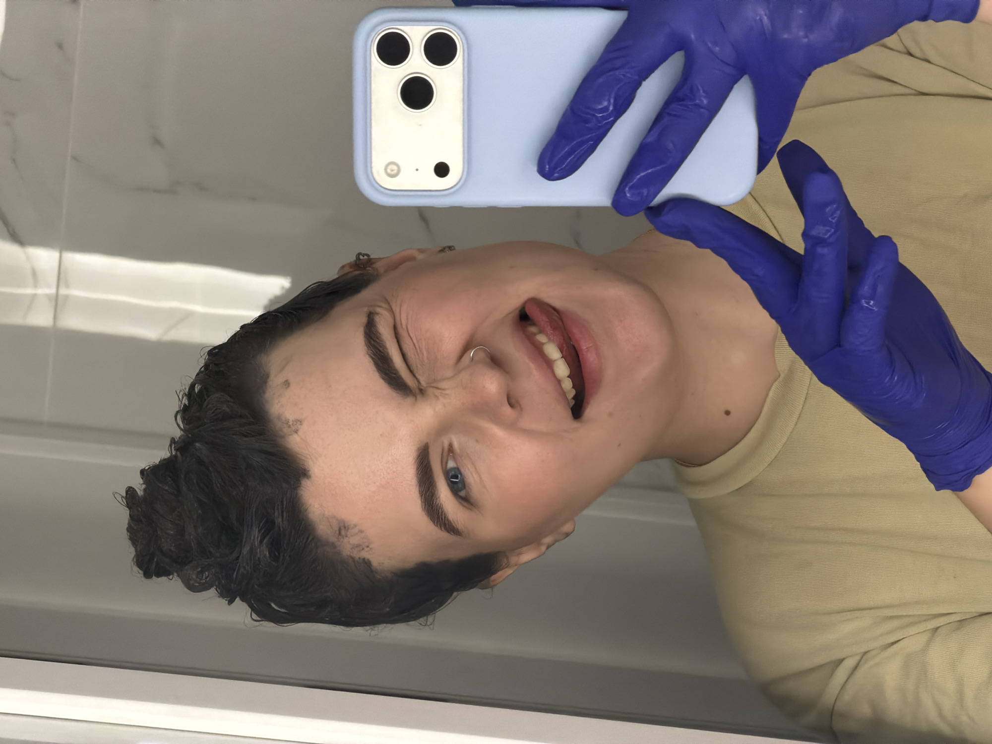 A selfie of a person with short, dark, wet hair and light skin tone, wearing a tan colored shirt and blue latex gloves. The person is holding a light blue smartphone case in their gloved hands. The individual is facing the camera at an angle, their face showing a playful expression with their tongue sticking out and one eye squinting. The background appears to be a bathroom with white tile or paneling. The location is not discernible from the image, and therefore cannot be placed in Chaikovsky, Russia.
