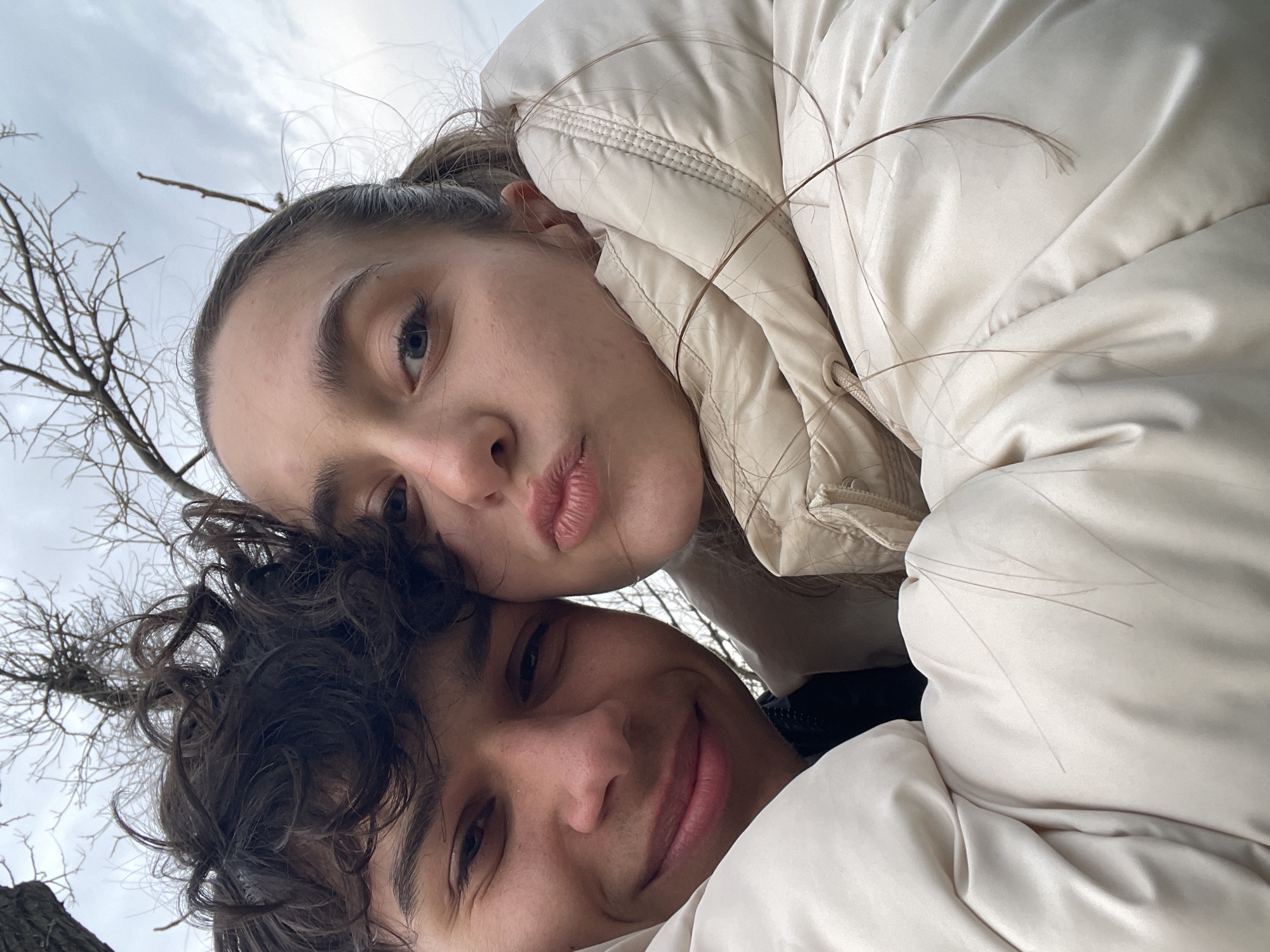Two individuals are pictured in a close-up, angled selfie. The person in the foreground has dark curly hair and is smiling slightly. The person in the background has fair skin, light brown hair pulled back, and is making a pursed-lip expression. Both are wearing light-colored puffy jackets. Bare tree branches are visible in the upper left of the frame. The location appears to be outdoors, potentially in a park in Varna, Bulgaria.