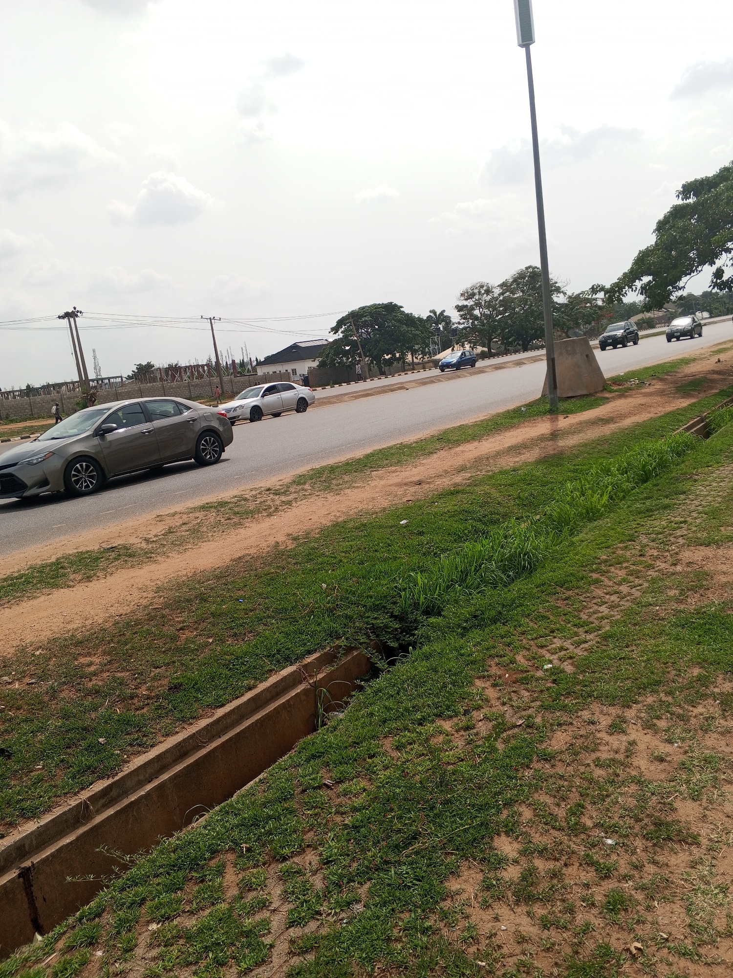 The image depicts a street scene in Abuja, Nigeria. A two-lane road curves slightly, with cars traveling in both directions. A dark gray sedan is in the foreground, moving left, and a light-colored sedan is directly behind it. Several other vehicles are visible further down the road.
A grassy area with a concrete drainage ditch runs along the right side of the road. Beyond the road, there is another grassy area with trees and buildings in the background. Overhead power lines are visible on the left side. A tall street lamp is on the right. The sky is overcast.