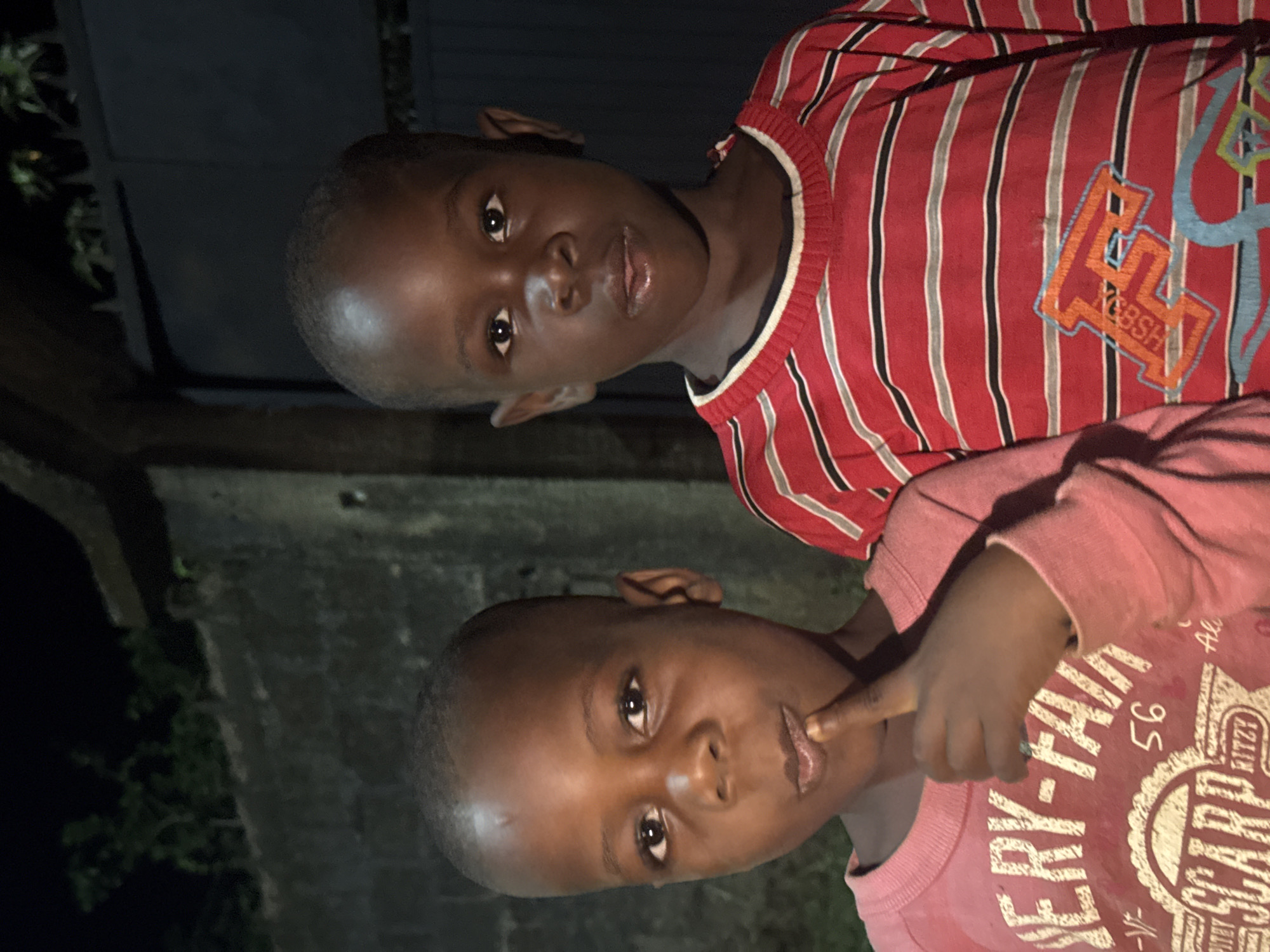 Two young children, appearing to be boys with dark skin, are captured in a close-up, dimly lit photograph. The image is oriented vertically, showing the children looking towards the camera.
The child in the upper portion of the frame wears a red long-sleeved shirt with thin black and white vertical stripes. An orange graphic design is visible on the chest, including letters resembling "E" and "SSH," possibly part of a brand name or logo. This child has a calm, direct gaze.
The child in the lower portion is wearing a pinkish-red long-sleeved shirt. White text and graphics are printed on it, with discernible words like "ERY-FAN", "56", "SCARF", and "RITZY". This child has their right index finger raised, lightly touching their mouth, a common gesture. Both children have short, dark hair and show highlights on their foreheads, suggesting direct frontal lighting, likely from a camera flash.
The background is dark, indicating it is likely nighttime or late evening. Behind the children, there appears to be a structure made of dark concrete blocks or bricks, with faint outlines of greenery or foliage visible on the far left. The setting is Eziudo, Nigeria.