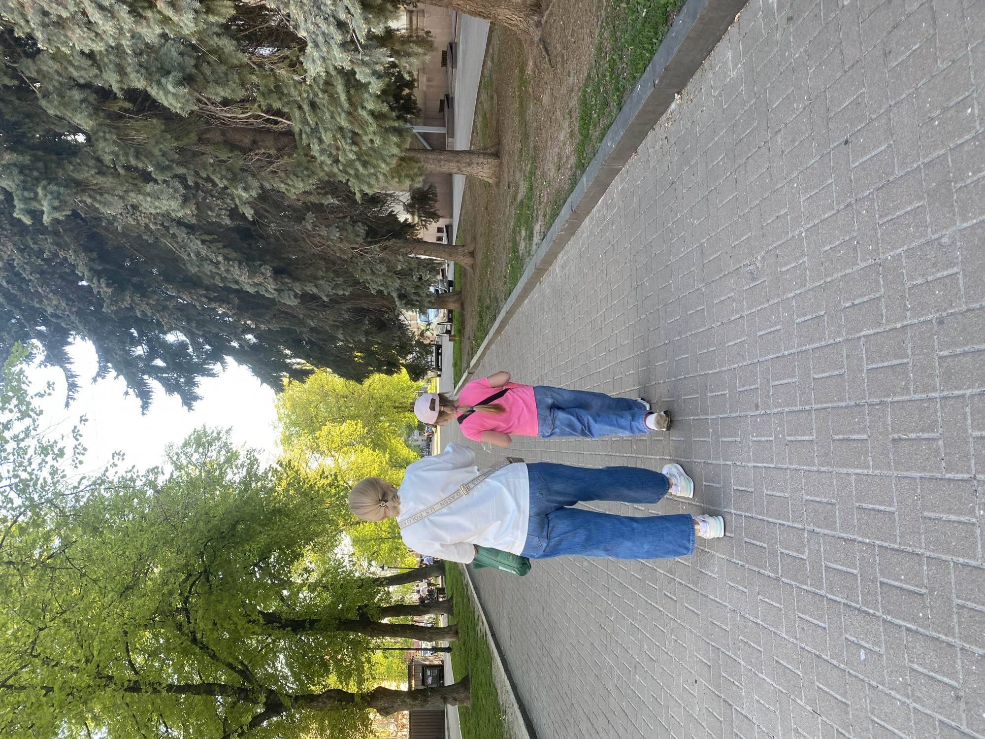 Two individuals walk side-by-side on a paved pedestrian walkway in the city of Poltava, Ukraine. The paved surface is made of rectangular blocks, with trees lining the left side of the walkway. The person closer to the camera wears a white top and blue jeans. The second person is wearing a pink shirt, blue jeans, and a pink hat. The environment suggests a park or public area.
