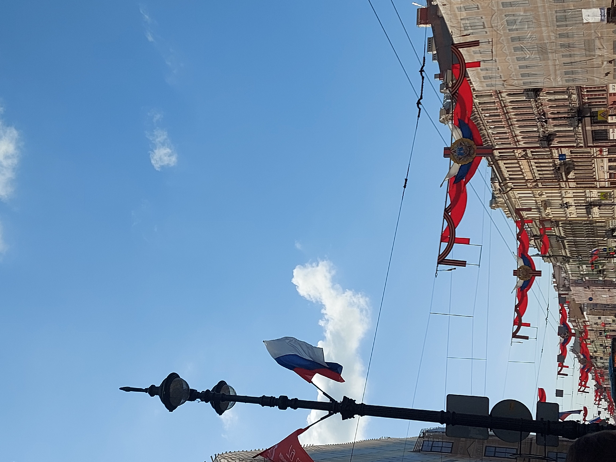 The image shows a view looking up at a street in Saint Petersburg, Russia, decorated with Russian flags and Victory Day symbols. The Russian tricolor flag is prominently displayed on a lamppost in the foreground. Red banners with Victory Day symbols are hung between buildings along the street. The sky is blue with scattered clouds. The buildings are light-colored and appear to be historic architectural structures. Streetlights are visible.