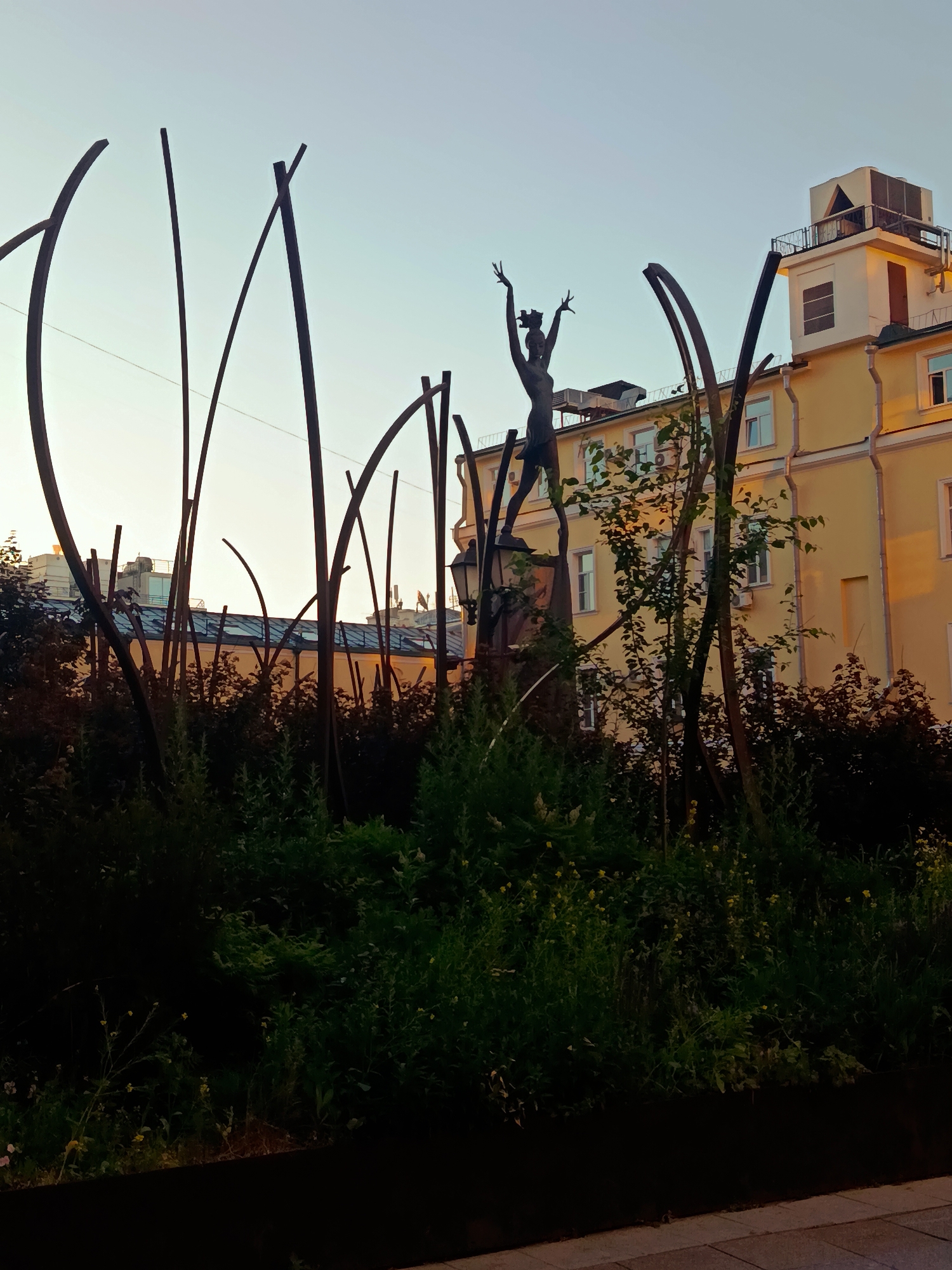 A low-angle shot captures a silhouette of a sculpture of a woman with her arms raised against a pale blue twilight sky. The sculpture stands amidst abstract, dark, curved metal elements and dense, dark green foliage in the foreground. Behind the sculpture, a bright yellow building with visible windows and rooftop air conditioning units is illuminated by warm light, suggesting late afternoon or early evening. The building appears to be a multi-story structure. The scene is set in an urban environment, with the building and the style of the sculpture indicating a city location. The overall impression is one of a public art installation within a city setting.