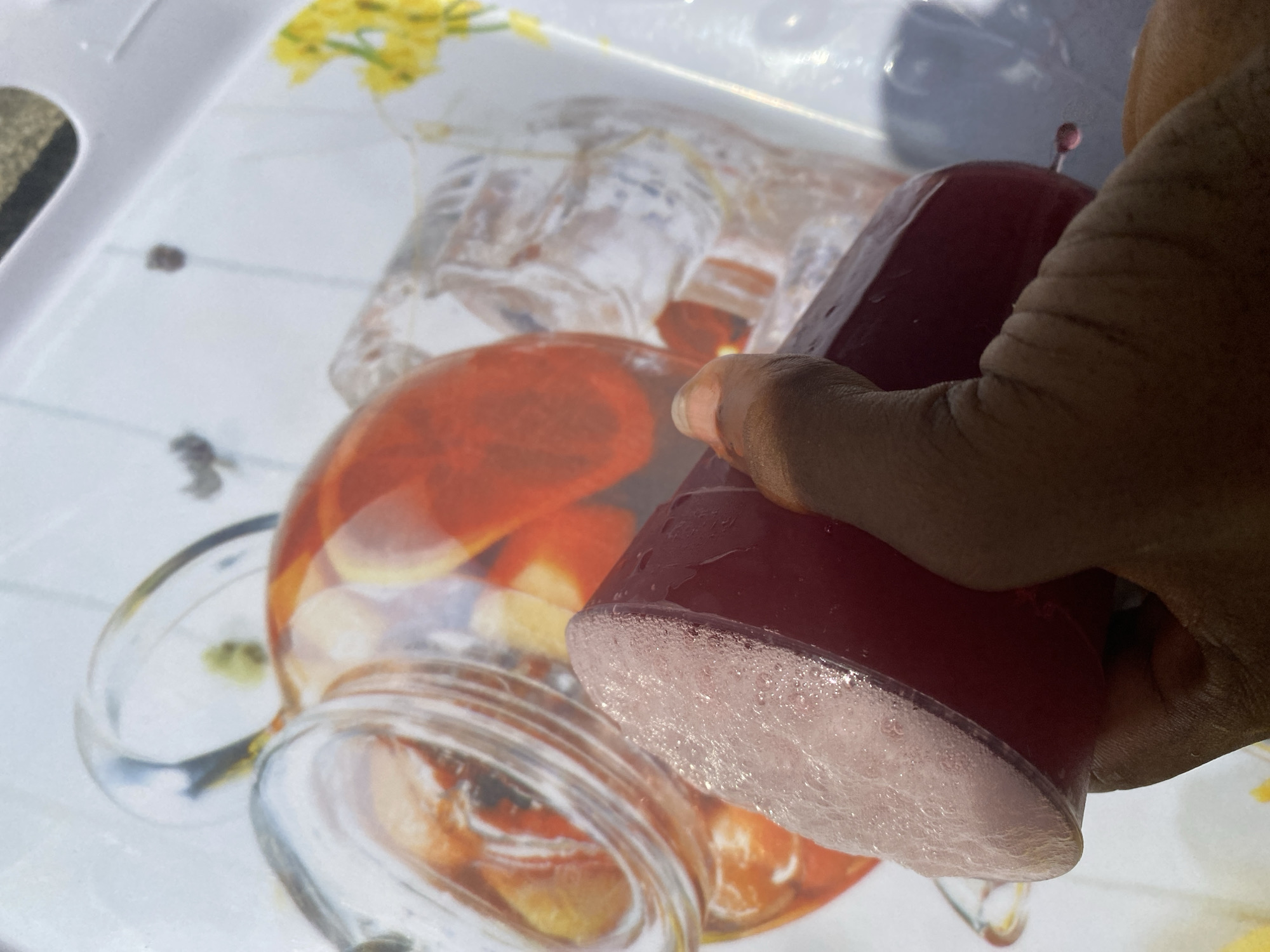 A dark-skinned hand holds a red, cylindrical candle with a light pink, textured bottom. The candle is positioned over a tray with a printed design featuring images of beverages in glass containers. The tray is white with a grid pattern. The environment appears to be outdoors, with bright light and shadows present. The specific location in the city of Iranla, Nigeria, cannot be determined from this image.