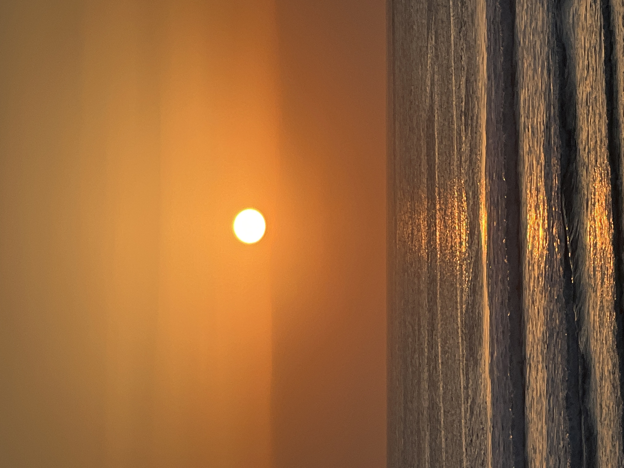A serene sunset over the ocean is captured in this vertical photograph. The sun, a bright white orb, is positioned slightly left of center in the upper half of the frame, casting a warm, orange glow across the sky and reflecting on the water. The ocean surface displays gentle waves and the shimmering path of sunlight. The image depicts the natural environment of Kuta, Indonesia, known for its beautiful beaches and vibrant sunsets, without any visible human presence or man-made structures.