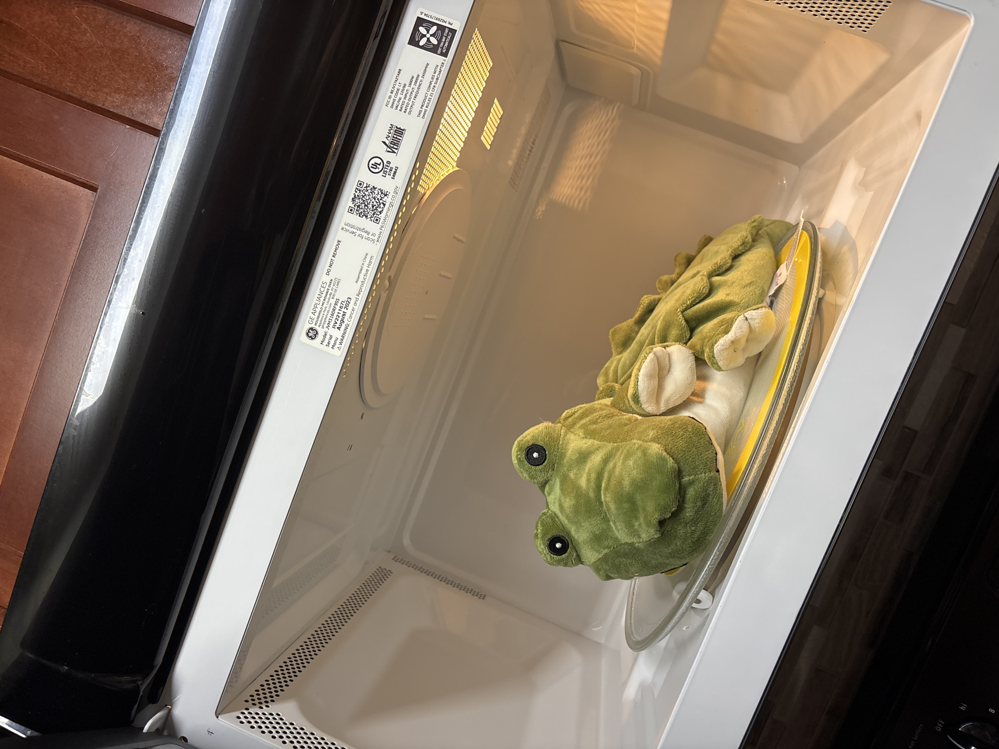 A plush alligator toy is resting on a glass plate inside a microwave oven. The microwave's interior light is on, illuminating the toy. The surrounding environment appears to be a kitchen, with wooden cabinets visible above the microwave. A label on the inside of the microwave door contains information about the appliance, including its model and serial number, and a warning about cancer and reproductive harm. The image was taken in Phoenix, Arizona, United States of America.