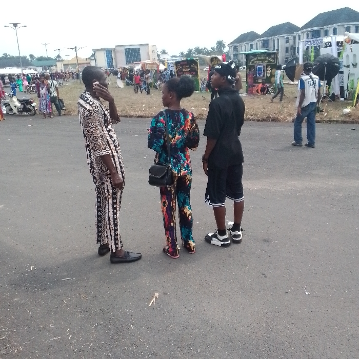 The image displays an outdoor scene in OGBOKO, Nigeria. In the foreground, three individuals stand on an asphalt surface. The leftmost individual, an adult male, wears a patterned black, white, and brown traditional two-piece outfit with black loafers, holding a light-colored mobile phone to his right ear. Adjacent to him, a young female with an afro hairstyle wears a multi-colored patterned top and trousers, a black shoulder bag, and pink-soled flat footwear, positioned with her back mostly to the camera. To her right, a young male wears a black t-shirt, black shorts with white trim, black and white sneakers, and a black baseball cap with white lettering, also facing away from the camera.
The mid-ground features a grassy area occupied by numerous people and various temporary structures or vendor stalls. One stall prominently displays "GOD IS GOOD" text. Two large black photographic umbrellas are visible on the right side of this area. A light-colored scooter is parked on the far left. In the distant background, several modern-style, multi-story buildings with dark roofs are visible under an overcast sky. Overhead streetlights are present on the left side of the frame.