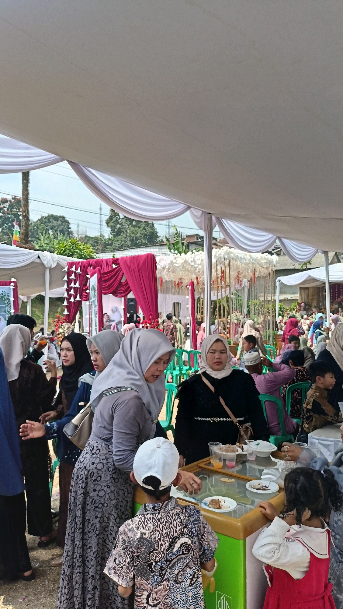 In Jambudipa, Indonesia, a large outdoor gathering is taking place under a white canopy, likely a wedding or festival. Many people are dressed in traditional Indonesian attire, with women wearing hijabs and colorful clothing. In the foreground, a woman in a grey hijab and patterned dress leans over a table, interacting with a young boy in a white cap and batik shirt. To the right, a young girl in a red dress stands by the table, looking at its contents. The table is covered with plates of food, drinks in cups, and other party supplies. The background reveals more attendees, decorations, and lush green trees, indicating a rural or semi-rural setting.