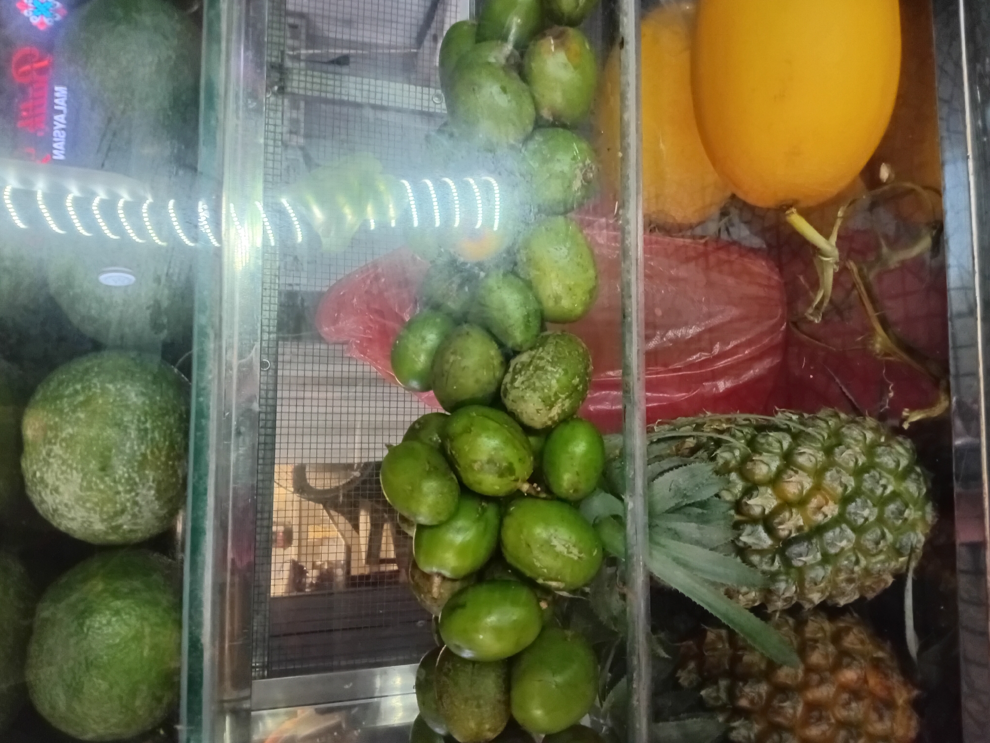 The image depicts a display of fresh fruits inside a glass-fronted refrigerator, likely a stall or shop in Đà Nẵng, Vietnam. The primary subjects are various fruits, including large green fruits that appear to be watermelons or melons, a pile of smaller green oval fruits resembling olives or small plums, two yellow fruits that could be papayas or mangoes, and two pineapples.
The fruits are arranged in sections within the refrigerator. In the leftmost section, several large, round green fruits are visible. The middle section showcases a cascade of smaller, green oval fruits. Behind these, red plastic bags are visible, hinting at packaging or other products. To the right, a large yellow fruit dominates the top portion, with another partially visible behind it. Below these yellow fruits are two pineapples, one more prominently displayed than the other.
The setting appears to be indoors, suggested by the artificial lighting, including a strip of white LED lights that create a bright, illuminated effect across the fruits. The background shows a grid-like structure, possibly part of the refrigerator's shelving or a cooling vent.
There is no clear indication of people or specific activities taking place, as the focus is solely on the fruit display. The time of day and weather are not discernible from the image.
Visible text includes "MAЯYAJAM" in red lettering, reflected on the glass of the leftmost section, likely from an external sign or advertisement. The overall impression is a vibrant and fresh selection of produce offered for sale.