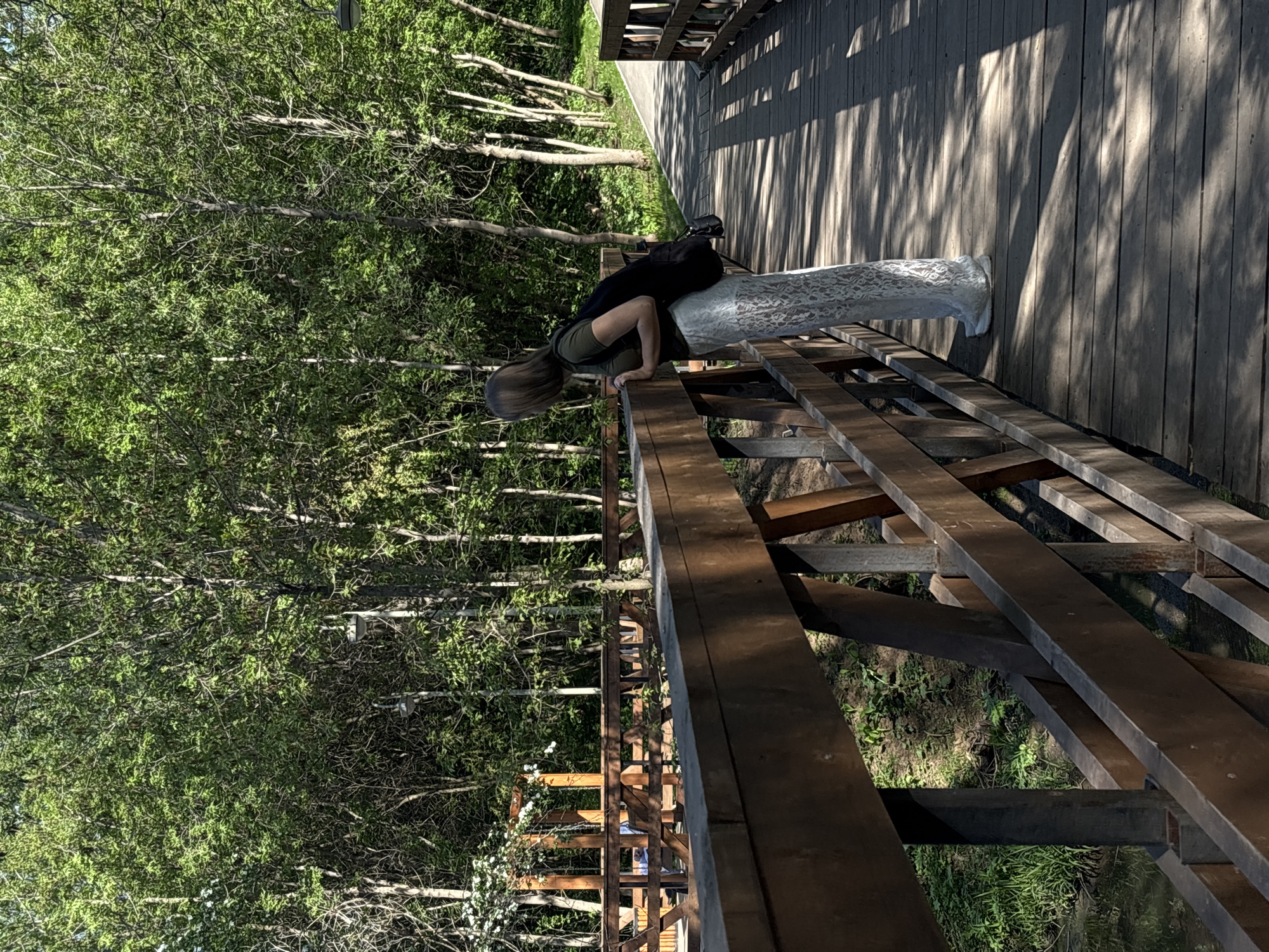 A woman is leaning over the railing of a wooden bridge, seemingly interacting with a cylindrical white object placed on the bridge's deck. The bridge structure is visible, with wooden planks forming the walkway and a wooden railing. Lush green foliage and trees are in the background. The bridge may be located in or near a wooded area. It could be located in the city of Perm, Russia.