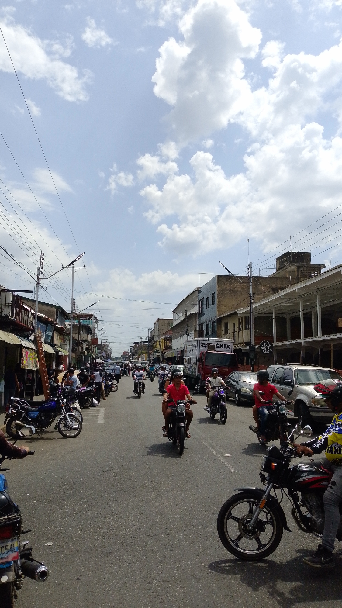 The image depicts a street scene in Tinaquillo, Venezuela. The scene is busy with traffic, particularly motorcycles, which are prominent in the foreground and middle ground. Several people are riding the motorcycles, some wearing helmets, and others not.
The buildings lining the street are a mix of one and two-story structures, some with businesses on the ground floor and apartments above. Some have balconies or awnings extending over the sidewalk. Power lines stretch across the sky above the street. A truck is parked on the side of the street. Cars and other vehicles are also present.
The weather appears to be sunny, with some clouds in the sky. The overall impression is of a bustling, everyday scene in a Latin American city.