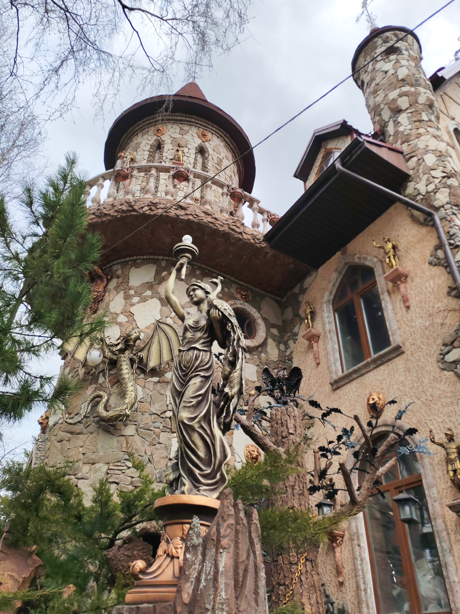 The image shows an elaborately decorated building resembling a fairytale castle in Izhevsk, Russia. The building features a stone tower with a reddish-brown conical roof, decorative balustrades, and stone detailing. A statue of a woman holding a torch stands in front of the tower. Other decorative elements include a dragon sculpture and various other embellishments on the facade. A second, taller stone tower with a cross on top is also visible on the right side of the image. The building is surrounded by greenery. The sky is overcast.
