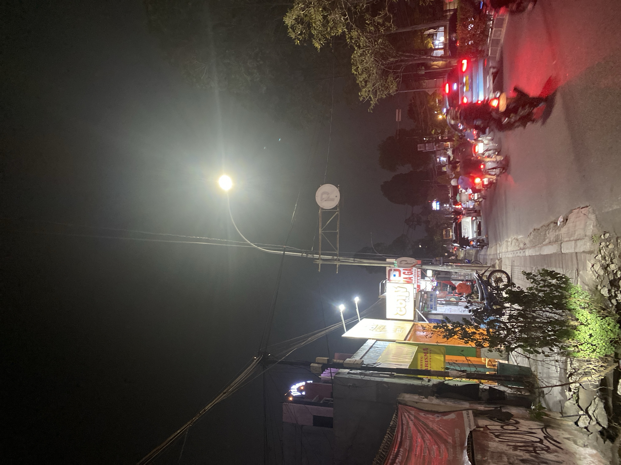 This is a nighttime, eye-level, full-shot photograph of a street scene in Tangerang, Indonesia. The image captures a busy road with numerous motorcycles and a few cars. A bright street lamp illuminates the center of the frame, creating lens flare. On the left side of the street, there are buildings with illuminated signs, including one that appears to say "MAGA" with "INDONESIA BANJAR" below it. A red banner is draped on a wall with graffiti. A sidewalk runs along the left side of the street, with some overgrown vegetation in the foreground. The road is filled with traffic, with motorcycles and cars moving in the same direction. The overall atmosphere is that of a bustling urban environment at night.