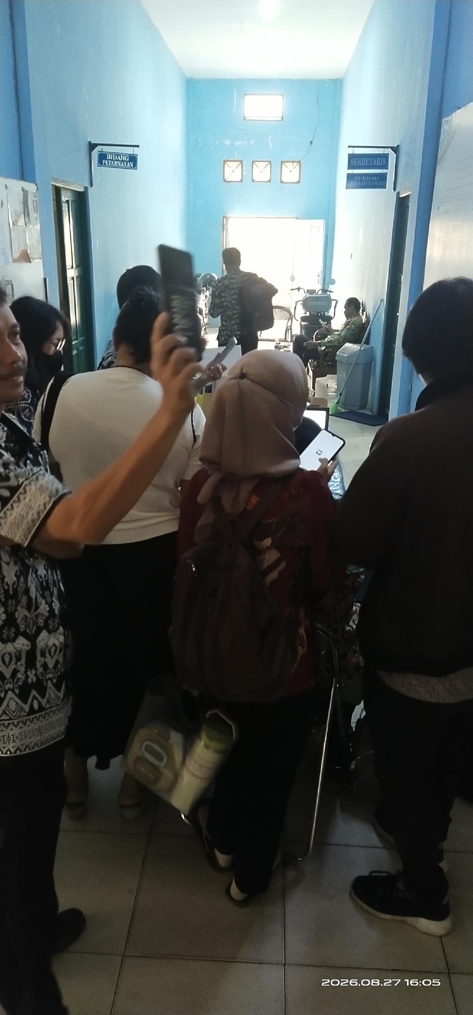 In Tamiang Layang, Indonesia, this image captures a hallway within a building, evidenced by the blue painted walls, tiled floor, and signage. Visible signs indicate "BIDANG PETERNAKAN" (Animal Husbandry Department) and "SEKRETARIS" (Secretary) with a sub-heading "SUBBAG TU & PERLENGKAPAN" (General Affairs and Supplies Sub-division). The hallway is populated by several individuals. In the foreground, a man on the left is partially visible, wearing a patterned shirt and holding a phone to take a picture or video. To his right, a woman in a white shirt and black pants is seen from the back, also holding a phone. Further into the hallway, another individual in a pinkish headscarf and dark patterned shirt with a backpack is looking at a phone. In the background, a person in a camouflage shirt is walking away, and another person in a military uniform is seated. Objects in the scene include chairs, a motorcycle, a trash can, and a clear plastic bag containing what appear to be food items and a bottle. The lighting suggests an indoor environment with natural light entering from the end of the hallway. The timestamp "2026.08.27 16:05" is visible in the bottom right corner.