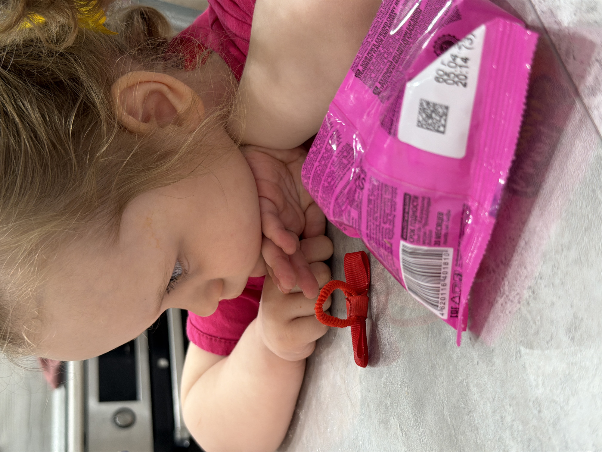 A fair-skinned child with blonde hair wearing a pink shirt is leaning on a surface next to a pink package. A small, red hair accessory is near the child's hand. The package shows a printed date of 05.04 20:14 and the barcode 4620116 401810. The product appears to be Sour Punch Extruded Chewy Jelly. The image has a domestic, indoor setting. The location "Chaikovsky, Russia" may be inferred from the presence of the Russian text and the specific barcode information.