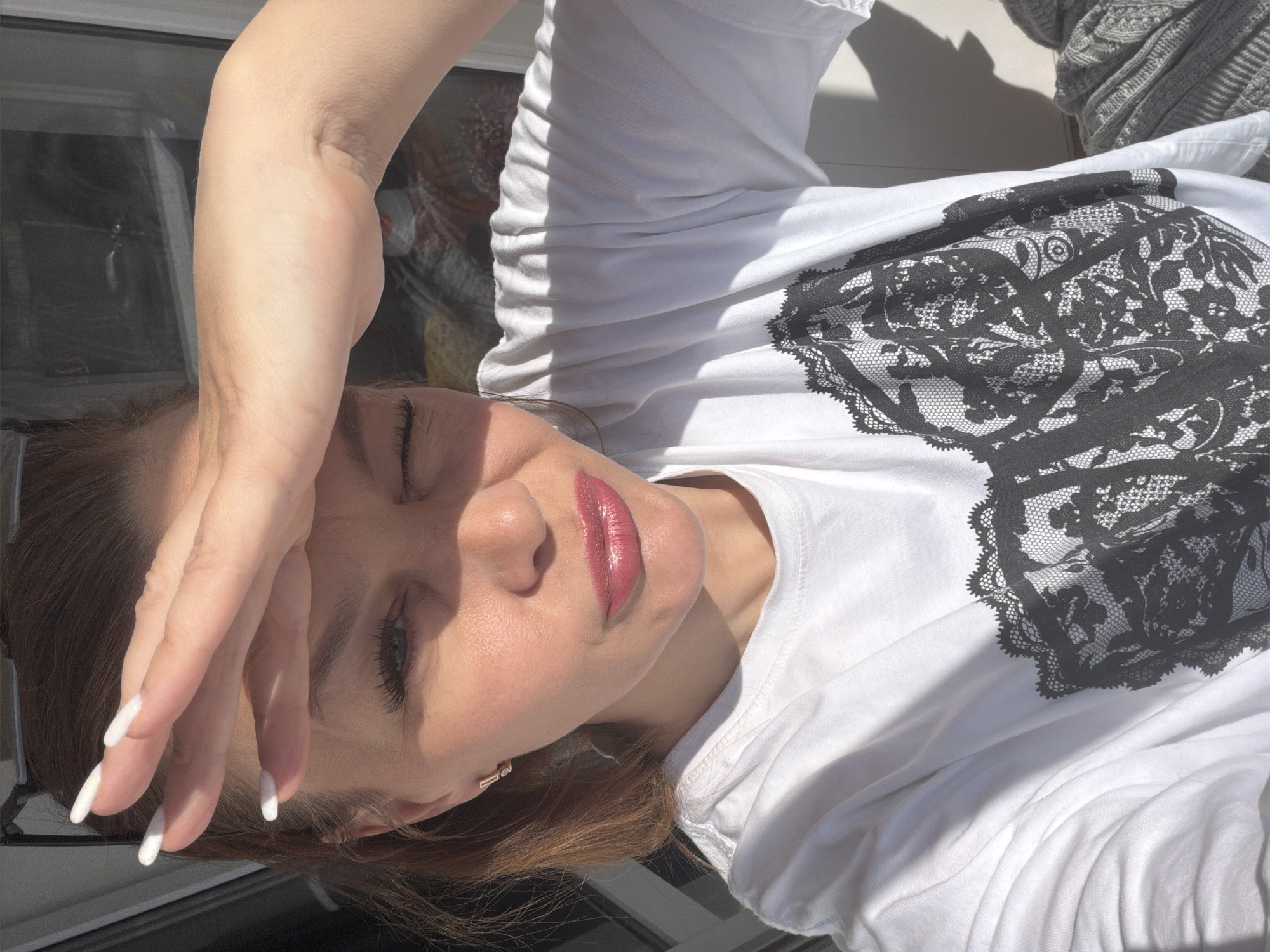A woman is posing for a selfie, shielding her eyes from the sun with her hand. She is wearing a white T-shirt with a black lace-like print on the front. She has manicured nails and is wearing lipstick. The background is blurred, but there is a window visible. The location is Chaikovsky, Russia.