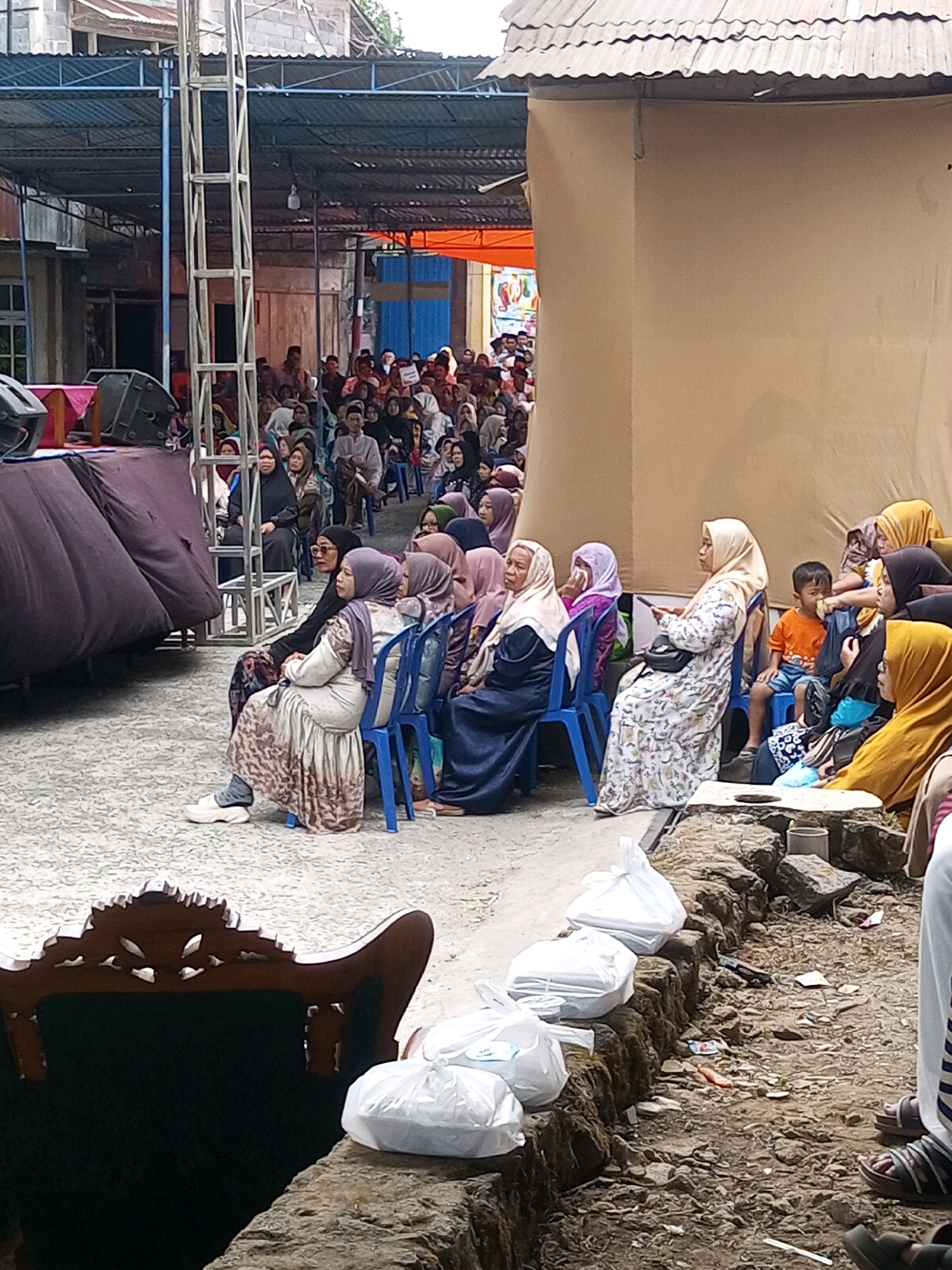 This image captures an outdoor gathering in Andongsili, Indonesia. A group of people, predominantly women wearing hijabs, are seated on blue plastic chairs, facing towards an unseen area, possibly a stage or presentation. The attendees are dressed in various traditional and modern clothing. Some individuals are interacting with their phones, while others appear to be engaged with the event. In the foreground, a carved wooden chair is partially visible, with white plastic bags and natural rocks forming a rough border. The background reveals a makeshift structure with a corrugated metal roof, suggesting a temporary setup for the event. The ground is a textured, greyish surface, likely asphalt or concrete, interspersed with dirt and debris. The overall scene depicts a community event or religious gathering taking place in a semi-urban or rural setting in Andongsili.