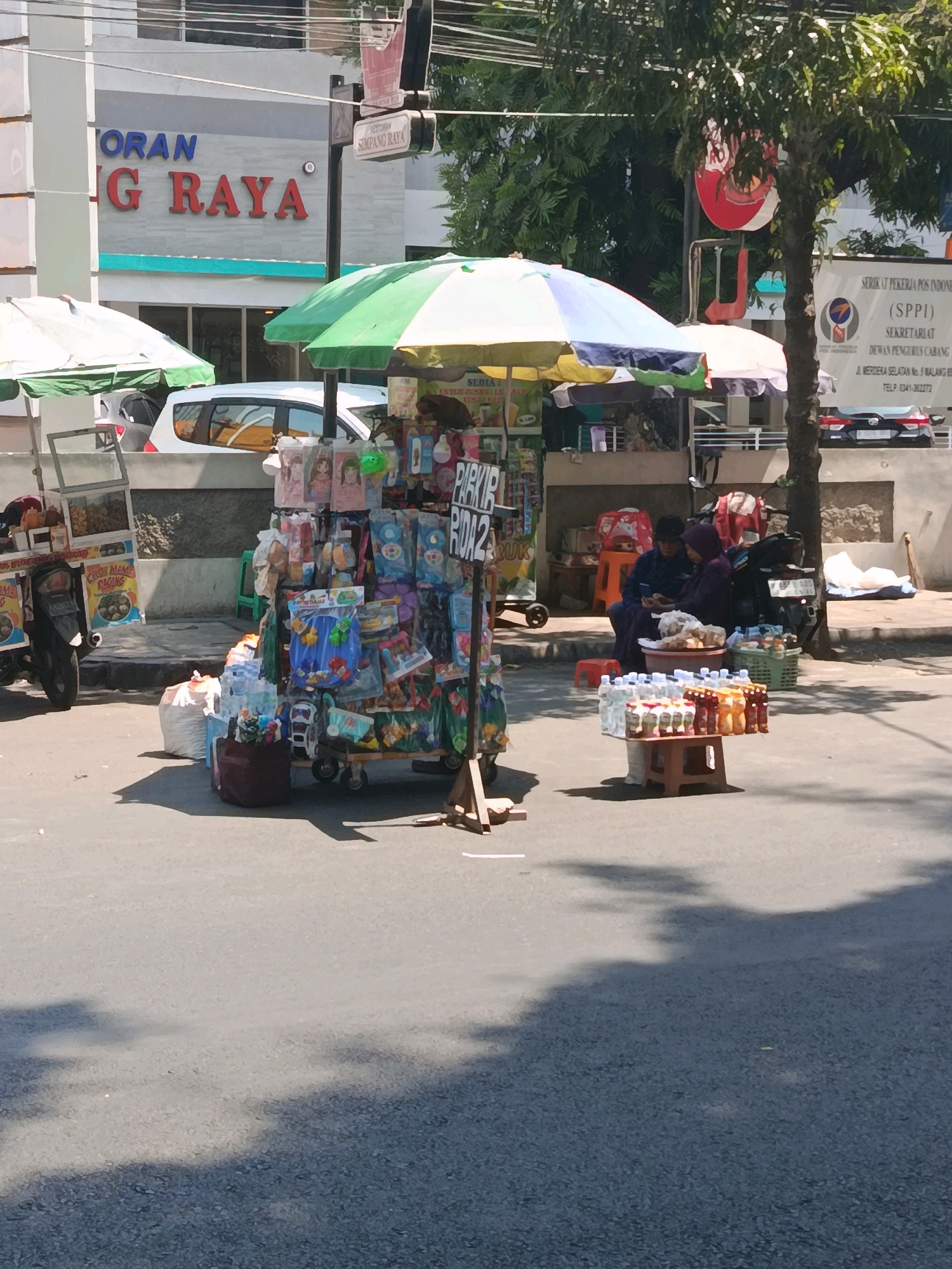 In Malang, Indonesia, a street vendor is operating on the side of a road, likely near a commercial area based on the building signage. The vendor has set up a stall under a green and white striped umbrella, displaying a variety of goods. To the left, a motorcycle-pulled cart is visible, also laden with items. In front of the stall, several plastic bags are stacked, and a collection of bottled drinks is displayed on a small wooden table. Two individuals, seemingly the vendors, are seated and appear to be interacting with each other. The environment is characterized by a paved road, concrete sidewalks, and a backdrop of buildings and trees under bright daylight. The overall scene depicts a typical street-level commercial activity in a bustling urban setting.