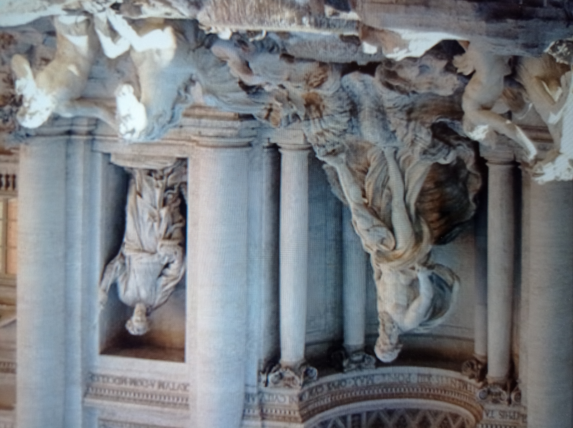 The image displays a portion of a large, ornate fountain, likely a famous landmark given the detailed stonework and sculpted figures. The style of the sculptures, particularly the classical attire and musculature of the figures, suggests a Baroque or Neoclassical influence. On the left, a large statue depicts a male figure with a beard, draped in flowing fabric, appearing to recline or struggle amongst rocks and water. To his right, another niche contains a standing female figure holding an object, possibly a staff or scepter. In the foreground, at the bottom right, two sculpted horses are shown, one rearing and the other partially submerged, with a human figure interacting with them. The overall scene is grand and dramatic, with a sense of movement and power conveyed through the dynamic poses of the figures and the representation of water. The environment appears to be an urban setting, with architectural elements like columns and decorative stonework forming the backdrop. The image quality is somewhat grainy, suggesting it may have been taken with a lower-resolution camera or under challenging lighting conditions. There is no visible indication or contextual information within the image that connects it to the city of Mukachevo, Ukraine.