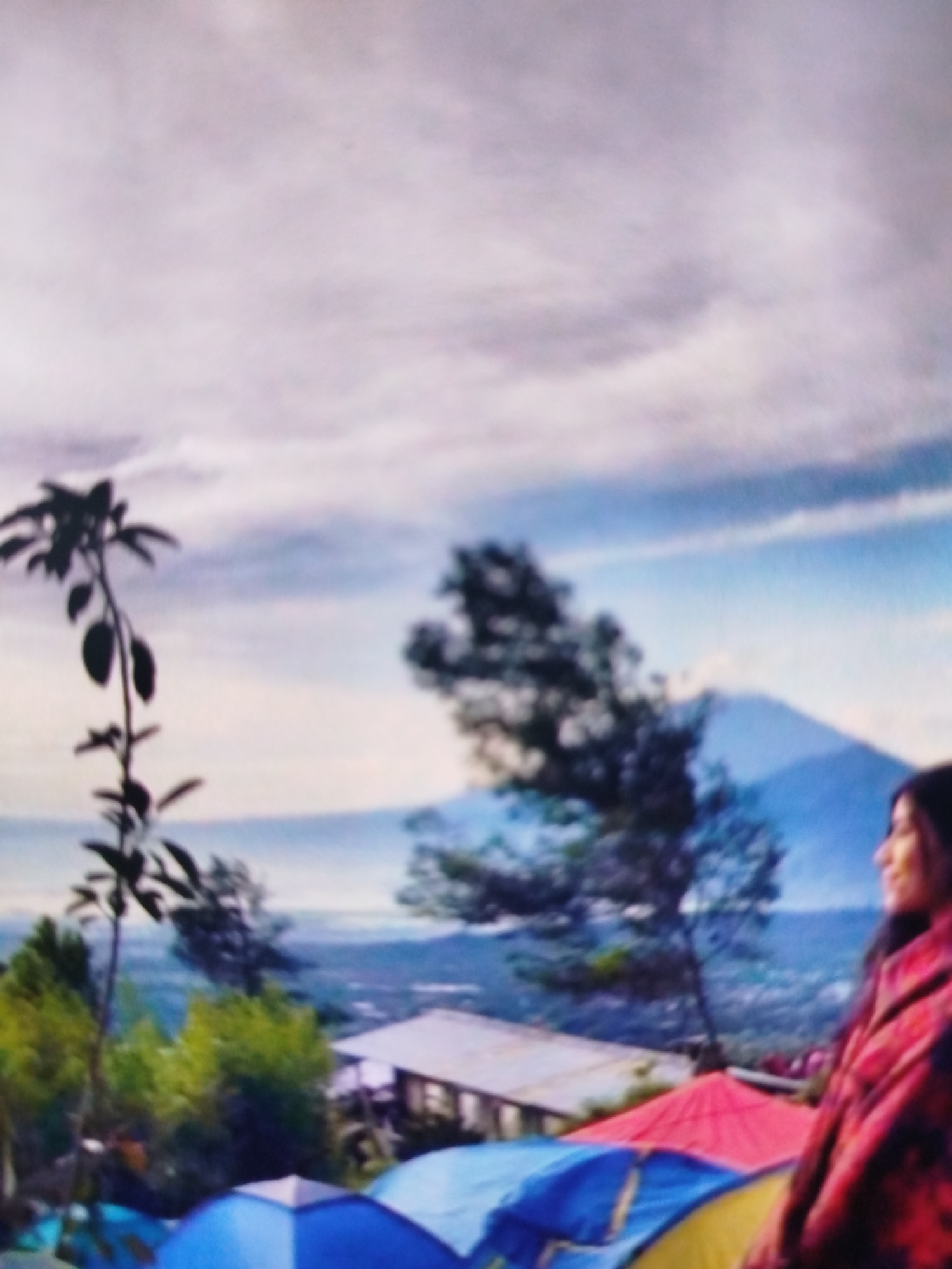 The image depicts a person on a hillside overlooking a valley and a distant mountain. The person, wearing a red and black patterned garment, is looking to the right. In the foreground, several tents are set up, appearing to be blue, yellow, and red. A building with a white roof is visible behind the tents. Lush green trees and a thin, dark-leaved plant are scattered across the hillside. The sky is cloudy with patches of blue. The scene suggests a camping or outdoor gathering setting on a natural elevation.