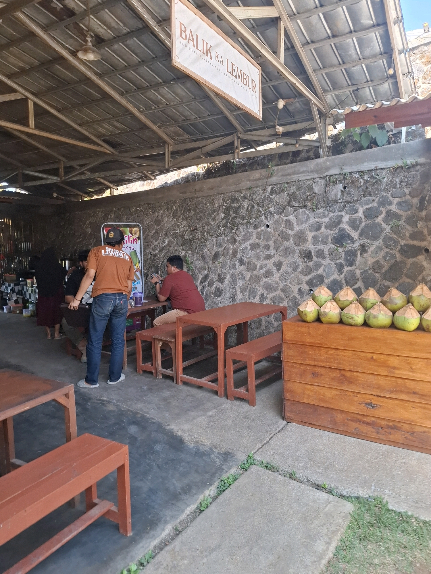 The image captures an outdoor seating area, likely a food stall or restaurant, situated in Cibentang, Indonesia. In the foreground, several empty wooden tables and benches, painted a reddish-brown, are visible on a concrete floor interspersed with patches of grass. To the right, a wooden cabinet filled with coconuts is prominently displayed. Behind this, a stone wall forms the backdrop, with a corrugated metal roof supported by a metal frame overhead, suggesting a rustic, open-air establishment.
In the mid-ground, a group of individuals are interacting around a table. A man wearing an orange t-shirt with "BALIK KA LEMBUR" printed on the back and a baseball cap is standing, facing away from the camera. He appears to be talking to another man seated at the table, who is wearing a maroon shirt and looking at his phone. Another person, possibly a woman in a dark headscarf and skirt, is standing to the left, partially obscured by the man in the orange shirt. A vertical display board with colorful images is positioned behind them.
The overall impression is of a casual, local gathering place. The presence of coconuts suggests a tropical setting, and the signage indicates a business catering to local patrons. The lighting suggests it is daytime, with some sunlight filtering through the roof.
