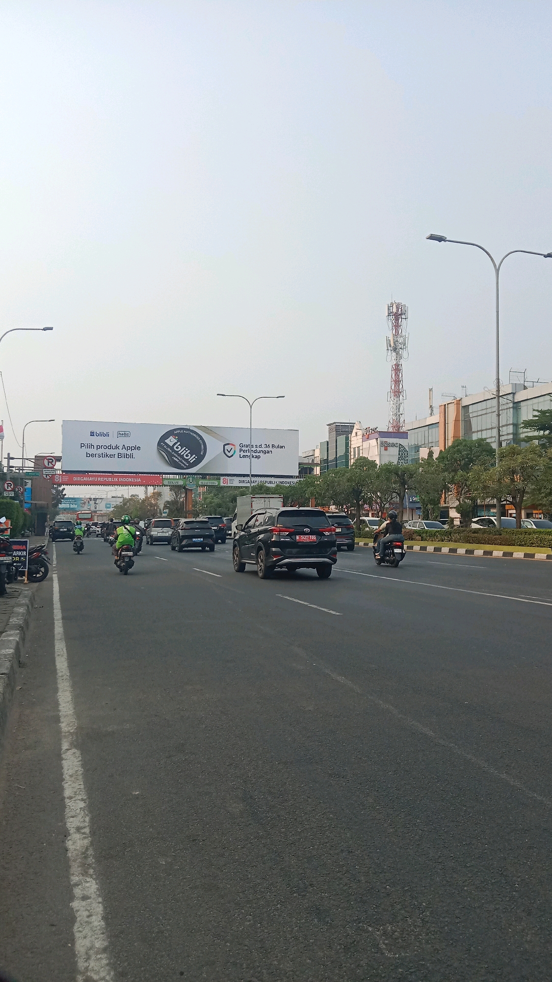 A street scene in Bekasi, Indonesia, shows heavy traffic on a multi-lane road. A billboard advertises "Blibli.com" with the slogan "Pilih produk Apple berstiker Blibli." Vehicles include several cars, some of which appear to be SUVs, and numerous motorcycles. Some motorcycles are carrying passengers, and several riders are wearing bright green vests, suggesting they might be delivery personnel. Buildings line the right side of the road, featuring modern architecture with glass facades and trees planted along the sidewalk. A tall cell phone tower is visible in the distance. The sky is overcast, casting a diffused light over the scene. The road is paved and appears to be in good condition, with clear lane markings. The overall impression is a busy urban environment during daylight hours.