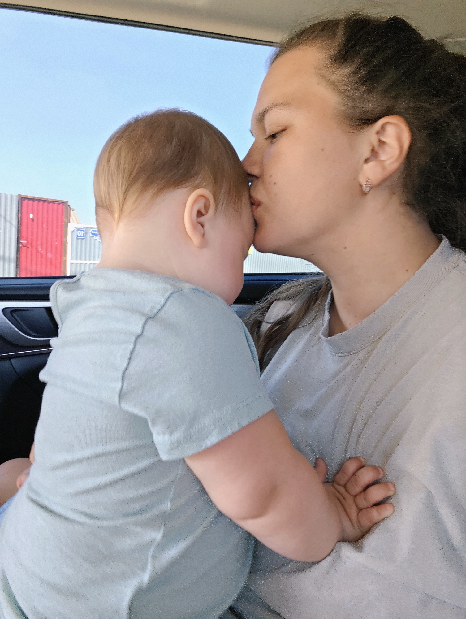 A woman with dark hair pulled back is kissing a baby with light hair on the forehead. Both are wearing light-colored t-shirts. They appear to be inside a car. Outside the car window are shipping containers and a blue sky. The sign on the containers indicates the Cyrillic letters "OR." This may indicate they are in the city of Malyi Bukor, Russia.