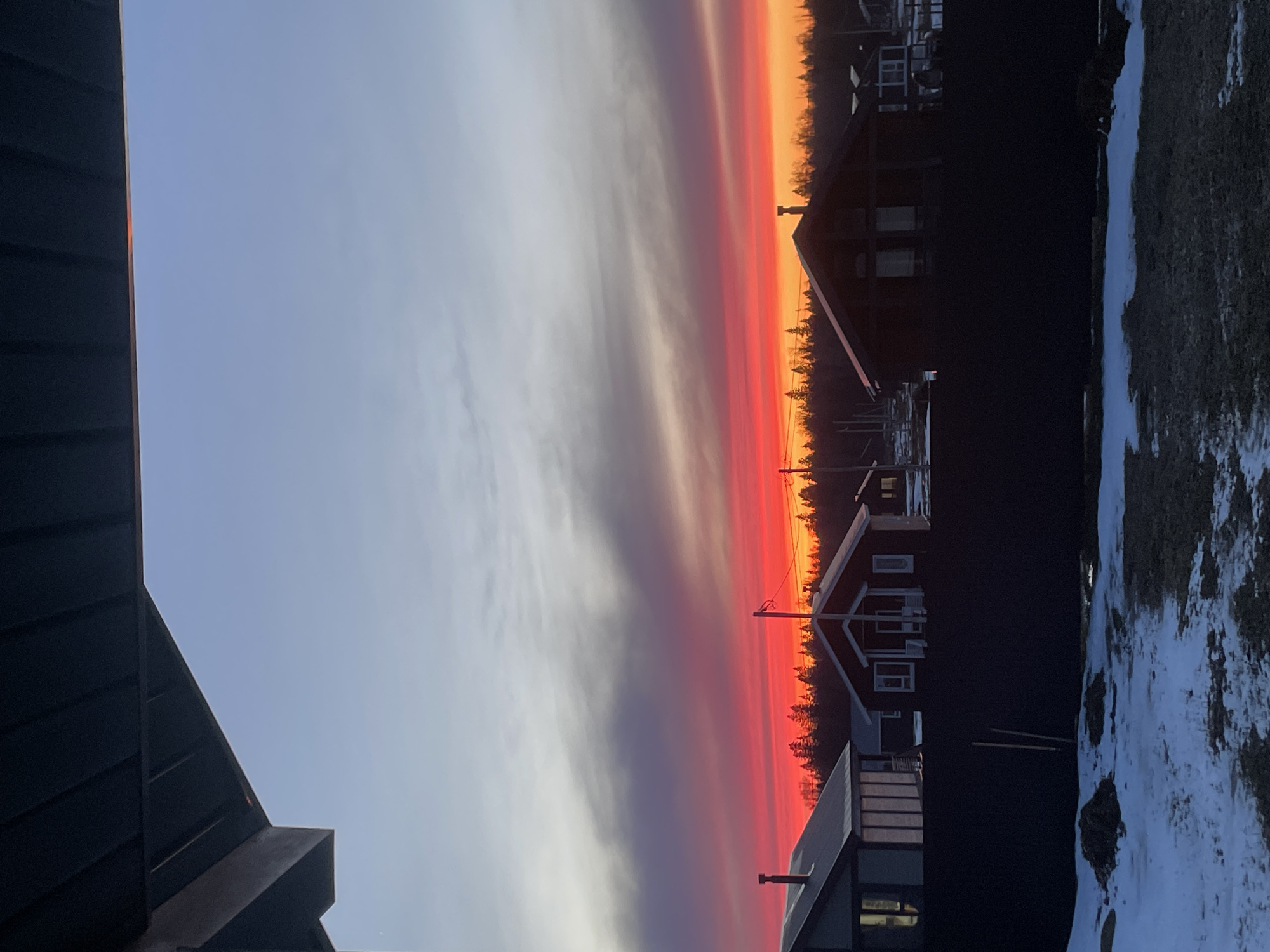 The image depicts a vibrant sunset scene with a clear division between a light blue sky on the left and a gradient of orange, red, and purple hues on the right, likely a sunset. Several houses are silhouetted against the colorful sky, suggesting a residential area. The scene also includes snow-covered ground, indicating a cold climate. The image appears to have been taken in Подшивалово, Russia, judging by the location information.