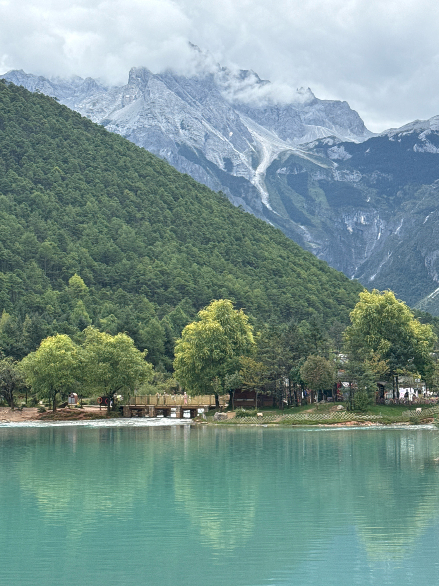 The image depicts a serene natural landscape in Yulong Nakhi Autonomous County, China. In the foreground, a tranquil, turquoise lake stretches across the frame, its surface rippling gently. The lake reflects the surrounding greenery and the overcast sky. Along the shoreline, a small wooden bridge connects two sections of land. Several trees with vibrant green foliage are scattered along the bank, some appearing to be willow trees due to their drooping branches. Behind the trees, a low fence encloses a grassy area where a few small buildings or stalls are visible. A few indistinct figures can be seen near the stalls and on the bridge, appearing to be tourists. The middle ground is dominated by a densely forested mountain slope, covered in a uniform green carpet of trees that ascends towards the background. The background features a majestic mountain range with rocky, snow-capped peaks partially obscured by clouds. The overall atmosphere is one of natural beauty and peacefulness, characteristic of a scenic tourist destination.