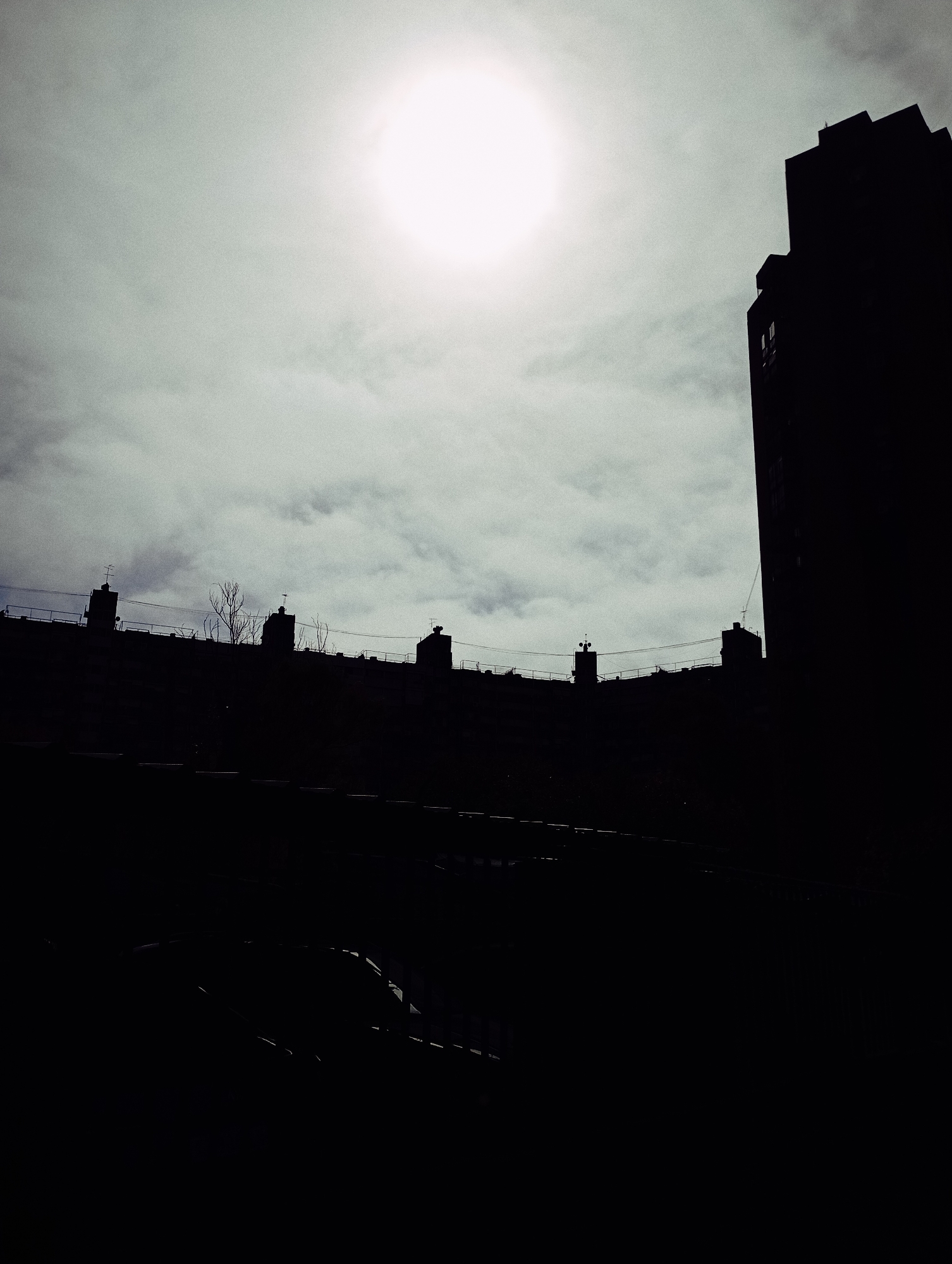 The image shows a silhouette of buildings against a bright sky in Buenos Aires, Argentina. The buildings have flat rooftops with various structures such as antennas, a bare tree, and chimneys. A taller building with visible windows dominates the right side of the frame. The foreground is mostly dark and obscured. The sky is cloudy, with the sun creating a bright halo.