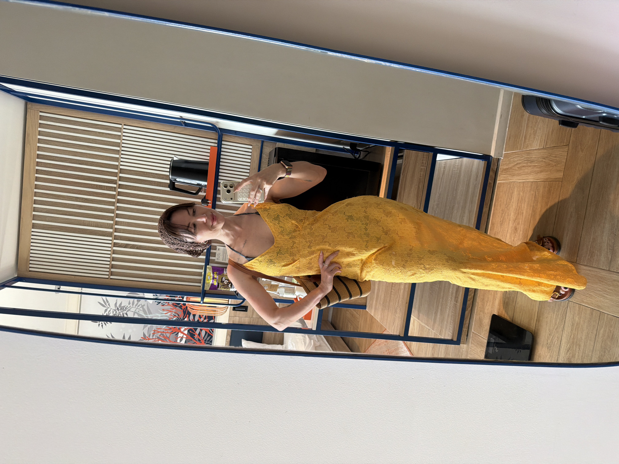 An adult female is depicted standing within a modern room, reflected in a full-length mirror, while taking a selfie with her smartphone. The woman, who has shoulder-length dark brown hair with bangs and is wearing a brown patterned headscarf, is smiling faintly while looking directly at the smartphone held in her left hand. A dark-banded smartwatch is visible on her left wrist, and a ring is on her left ring finger. She is dressed in a bright yellow, V-neck, sleeveless, full-length slip dress crafted from a textured, possibly lace or crocheted, semi-translucent fabric. Beneath the yellow dress, a dark blue and white patterned halter-style top is visible. Her right hand is positioned on her hip, holding a woven straw shoulder bag with vertical brown and black stripes. She wears brown open-toed sandals with a metallic buckle detail.
The room's interior, visible in the mirror's reflection, features light-toned wooden parquet flooring arranged in a diagonal pattern. A prominent feature in the background is a wall or partition designed with alternating vertical light wood and white striped panels. Adjacent to this, to the right of the woman's reflection, stands an open shelving unit constructed with blue metal frames and wooden shelves. The top shelf holds a black electric kettle resting on an orange tray. Lower shelves display items such as bottled water, a gold-colored snack bag, and a black electronic device, possibly a mini-fridge or speaker. Further left in the reflection, through an additional mirrored or glass panel, a wall with a graphic design of stylized red coral branches against a grey and white botanical pattern is visible. A white-sheeted bed is faintly discernible in a deeper reflection on the far left of the image. On the floor in the reflection, a black digital scale is located on the lower left, and a dark-colored suitcase is partially visible on the lower right. The scene is located in Dong Ninh Hoa Ward, Vietnam.
