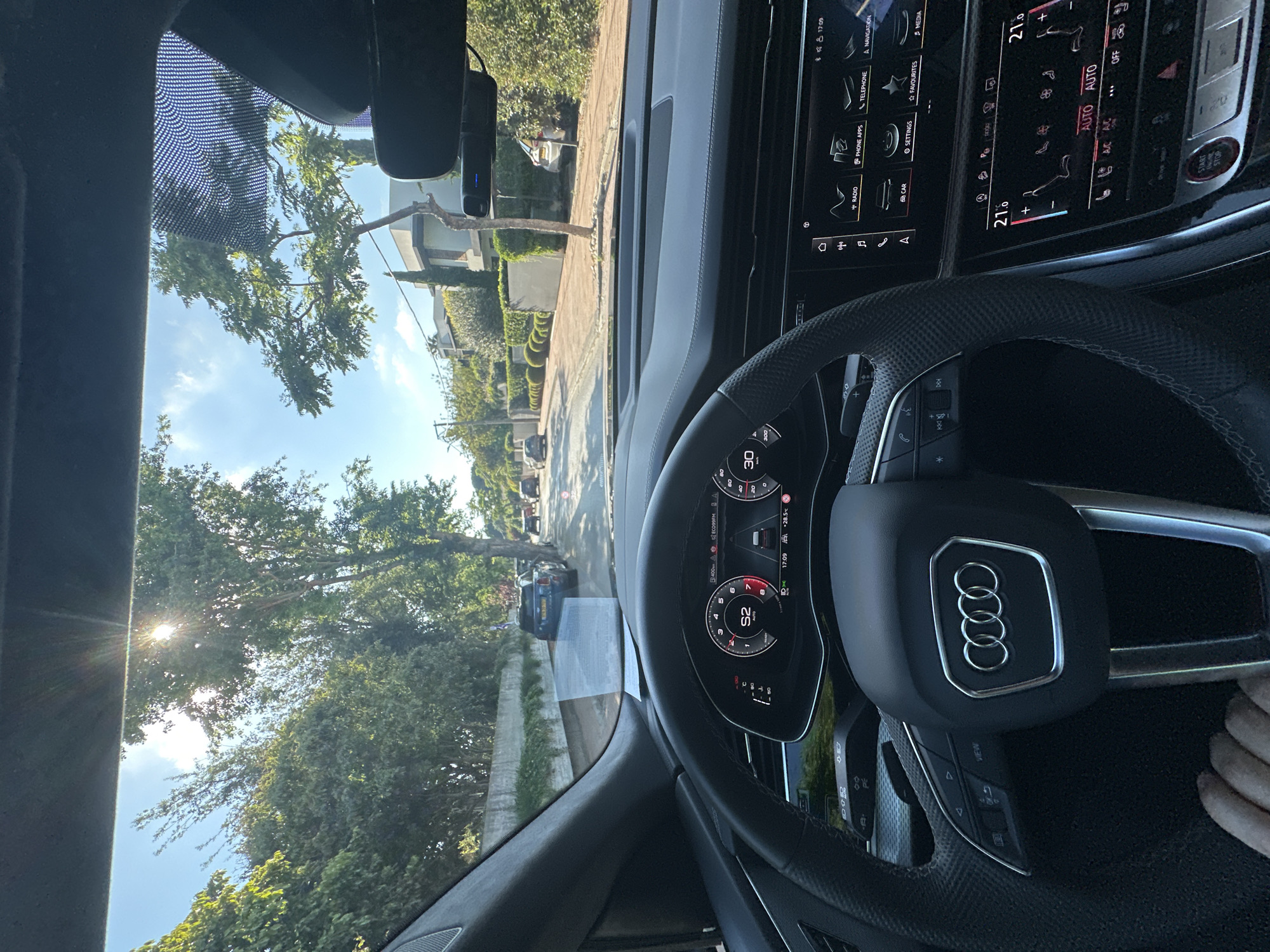 Captured from the driver's seat of an Audi, the image offers a perspective of the residential neighborhood of Kfar Shmaryahu, Israel. The car's dashboard is prominently featured, showcasing the steering wheel with the Audi emblem, digital instrument cluster displaying a speed of 52, and the infotainment system. The climate control settings are visible on the dashboard, set to automatic.
Outside the vehicle, a residential street is lined with trees and houses, typical of Kfar Shmaryahu. A car is visible in the distance. The time displayed on the car's digital instruments is 17:09. The sun is visible through the trees, causing lens flare. A portion of a person's fingers are visible in the bottom right corner of the image.