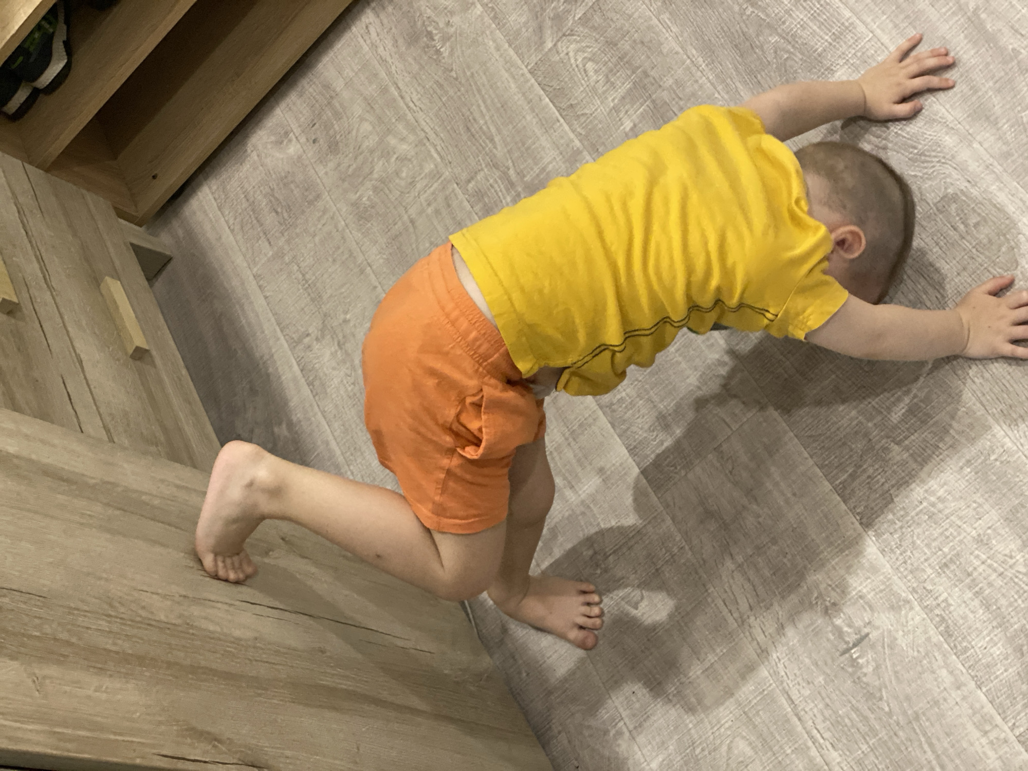 A young child is lying prone on a light-colored, wood-grain floor. The child is wearing a yellow t-shirt and orange shorts. The child's head is turned down and to the side, with the hands outstretched in front. The child's bare feet are visible, bent at the knees. Part of a light wood-colored cabinet is visible in the background. The location in the city of Sochi, Russia is not discernable from this image.