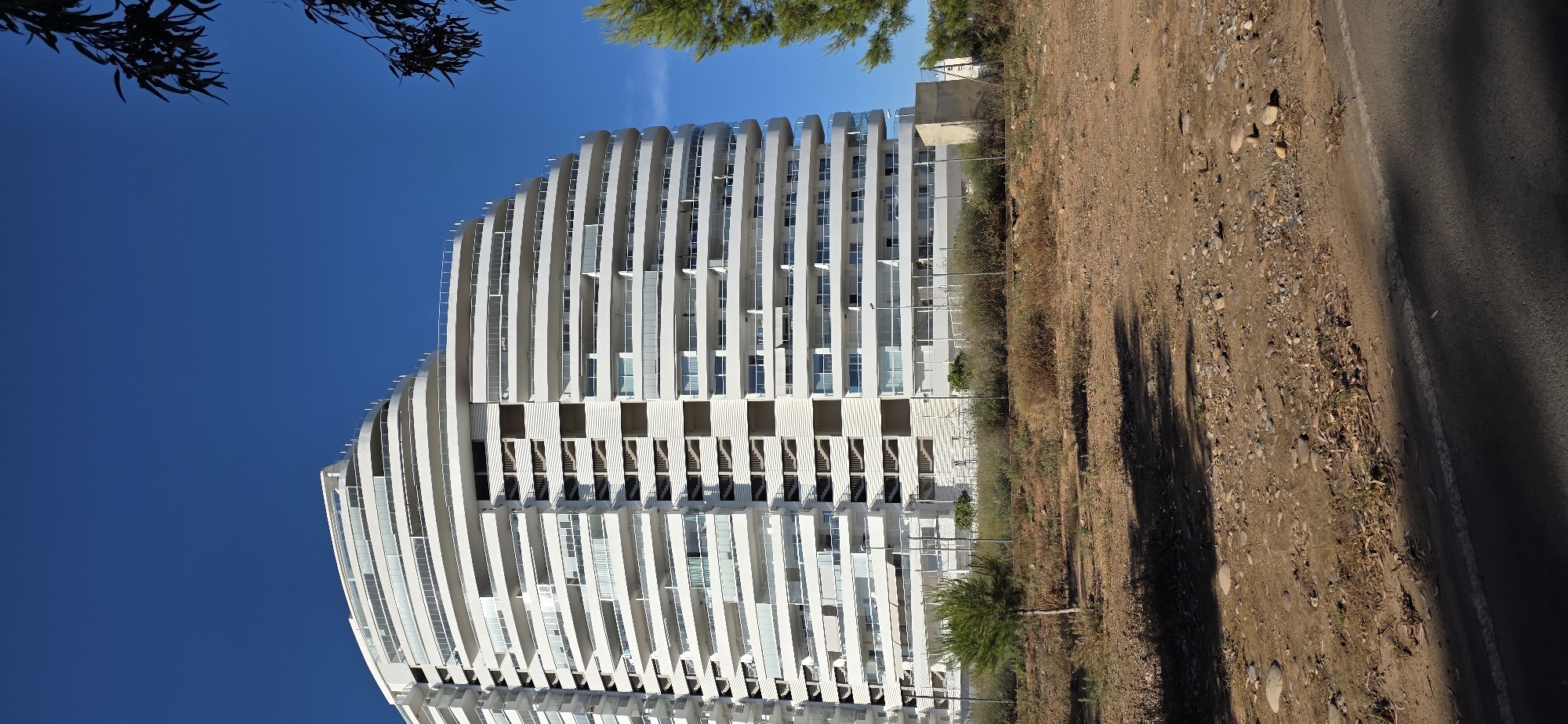 The image shows a tall, white, curved building in Canet d'en Berenguer, Spain. The building has many floors with balconies. To the right of the building is a dirt area, followed by a road with a white line. The sky is blue, and there are tree branches visible at the top of the image.