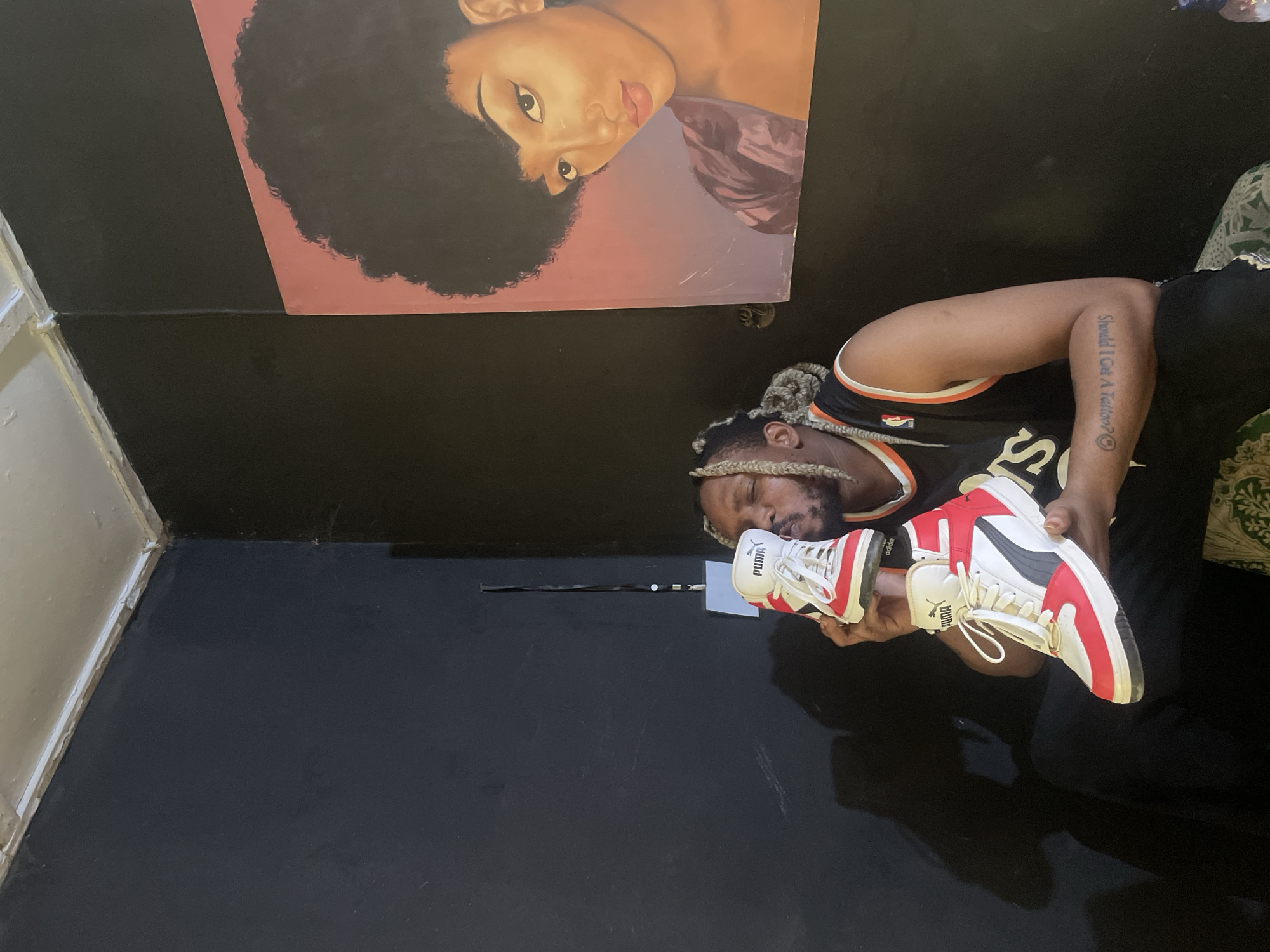 The image shows a man in a black basketball jersey holding a pair of red and white Puma high-top sneakers. The man has dreadlocks and a tattoo on his left arm that reads "Should I Get A Tattoo? :)". Above him is a painting of a woman with an afro. The background is a black wall and floor. There is no conclusive evidence within the image to confirm the image's location is Jos, Nigeria.