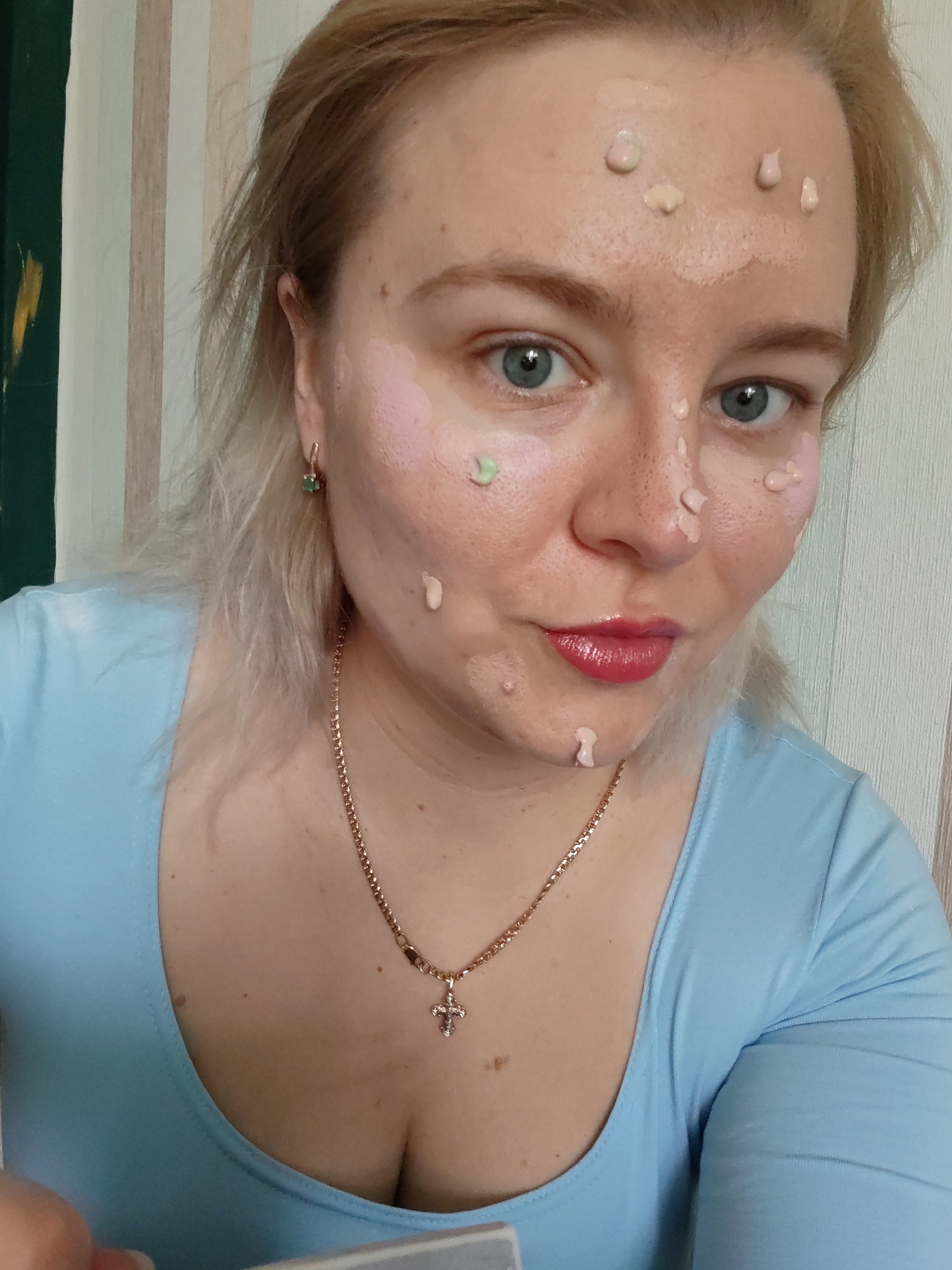 A fair-skinned adult female with blonde hair and light-colored eyes looks directly at the camera. Her face is dotted with multiple small dollops of cosmetic products in shades of beige, pink, white, and light green, suggesting an in-progress application. Her lips are coated with glossy red lipstick. She wears a light blue long-sleeved top with a scoop neckline. A gold-colored chain necklace with an ornate crucifix pendant hangs around her neck, and a gold-colored earring with a green stone is visible in her right ear. The background features a vertically striped wall in shades of light beige and white. To the far left, a dark green panel with a yellow mark is visible. A light-colored object is partially visible in the bottom left, held by the subject's hand.