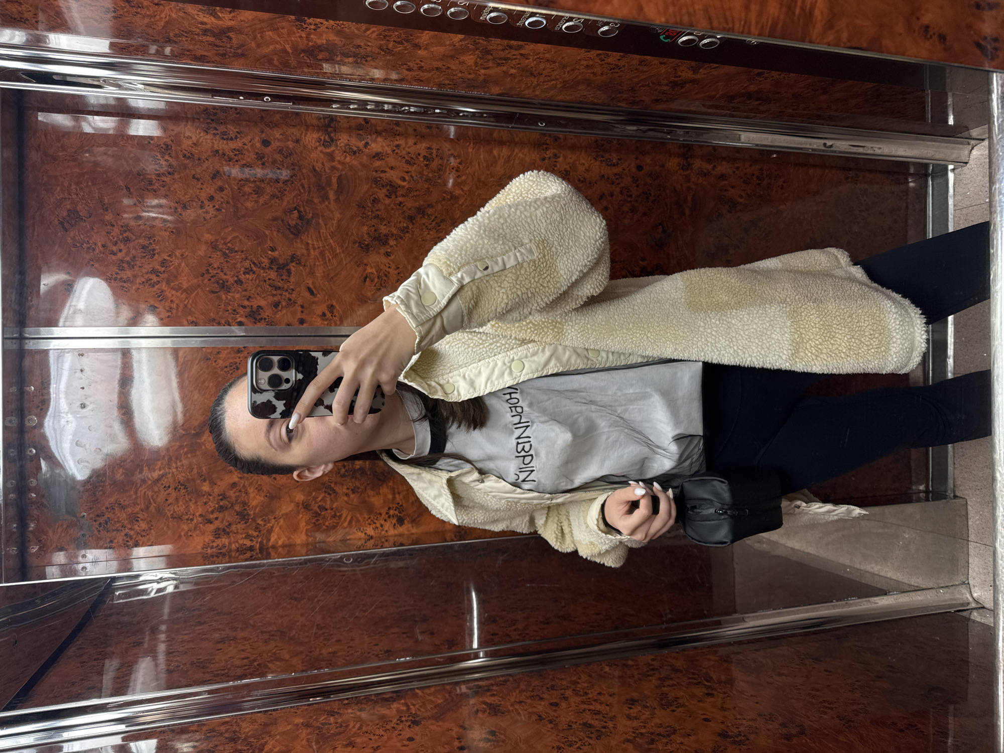 A woman is taking a selfie in an elevator with wood-paneled walls and a mirrored ceiling. She is wearing black leggings, a light gray t-shirt with writing, and a cream-colored, fleece-like jacket. She is holding a cell phone with a cow-print case. The environment is a modern elevator with metallic trim. The location is not discernible.