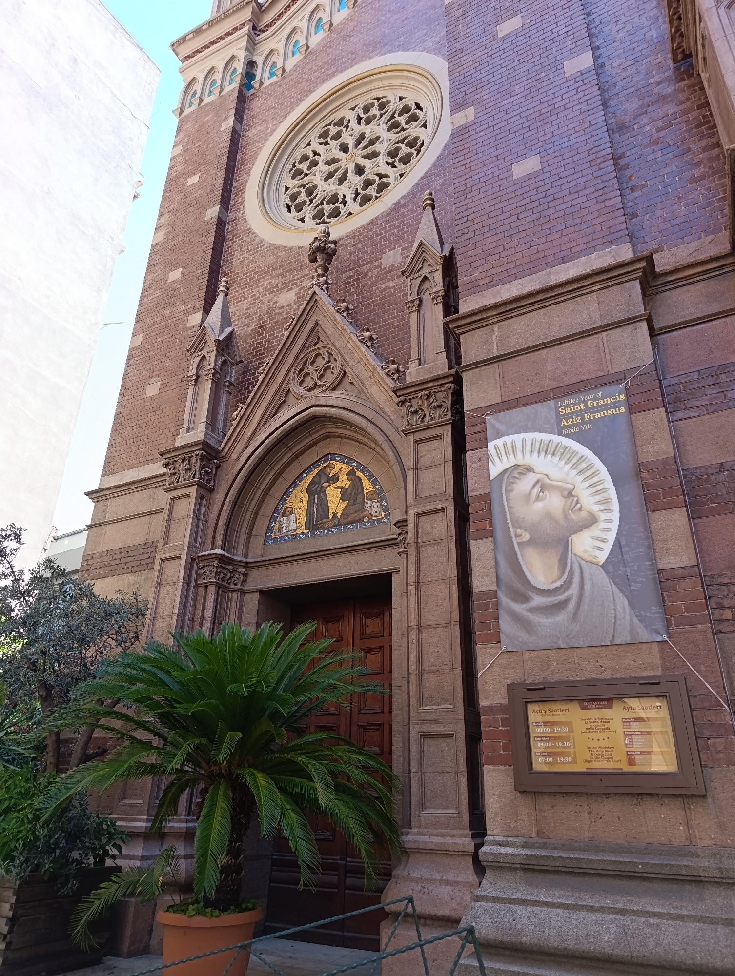 This is a low-angle, full outdoor shot of the exterior of the Saint Francis Church in Beyoğlu, Turkey. The church is constructed of red brick and features Gothic architectural elements, including pointed arches and a rose window above the main entrance. A large banner depicting Saint Francis of Assisi hangs on the right side of the facade. Below the banner is a brown informational sign displaying church hours in Turkish and English. In front of the church, a potted palm tree and other greenery are visible. The scene is brightly lit by daylight.