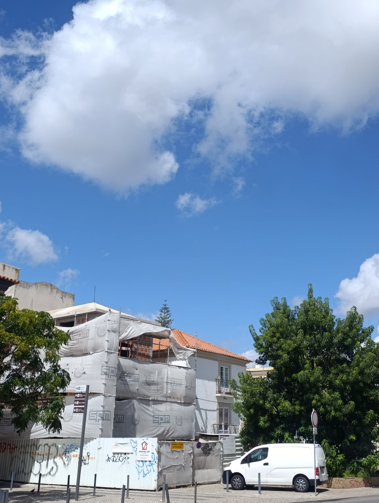 This is a low-angle, exterior, daytime, full-shot photograph taken on a bright, sunny day in Setúbal, Portugal. The sky is a vibrant blue with scattered white cumulus clouds. The foreground is dominated by a building under construction, draped in white protective sheeting. The sheeting is printed with a repeating logo that appears to be "SPM" and "CONSTRUÇÃO E REABILITAÇÃO". Some sections of the building's brickwork are visible through openings in the sheeting. A large, leafy green tree is situated to the right of the building. In front of the building, a white van is parked on the street. A traffic sign is positioned near the van. The street pavement is visible. Further to the left, the corner of another building with an orange tiled roof is visible, along with another large tree. The overall scene depicts an urban environment with ongoing construction and everyday life elements.